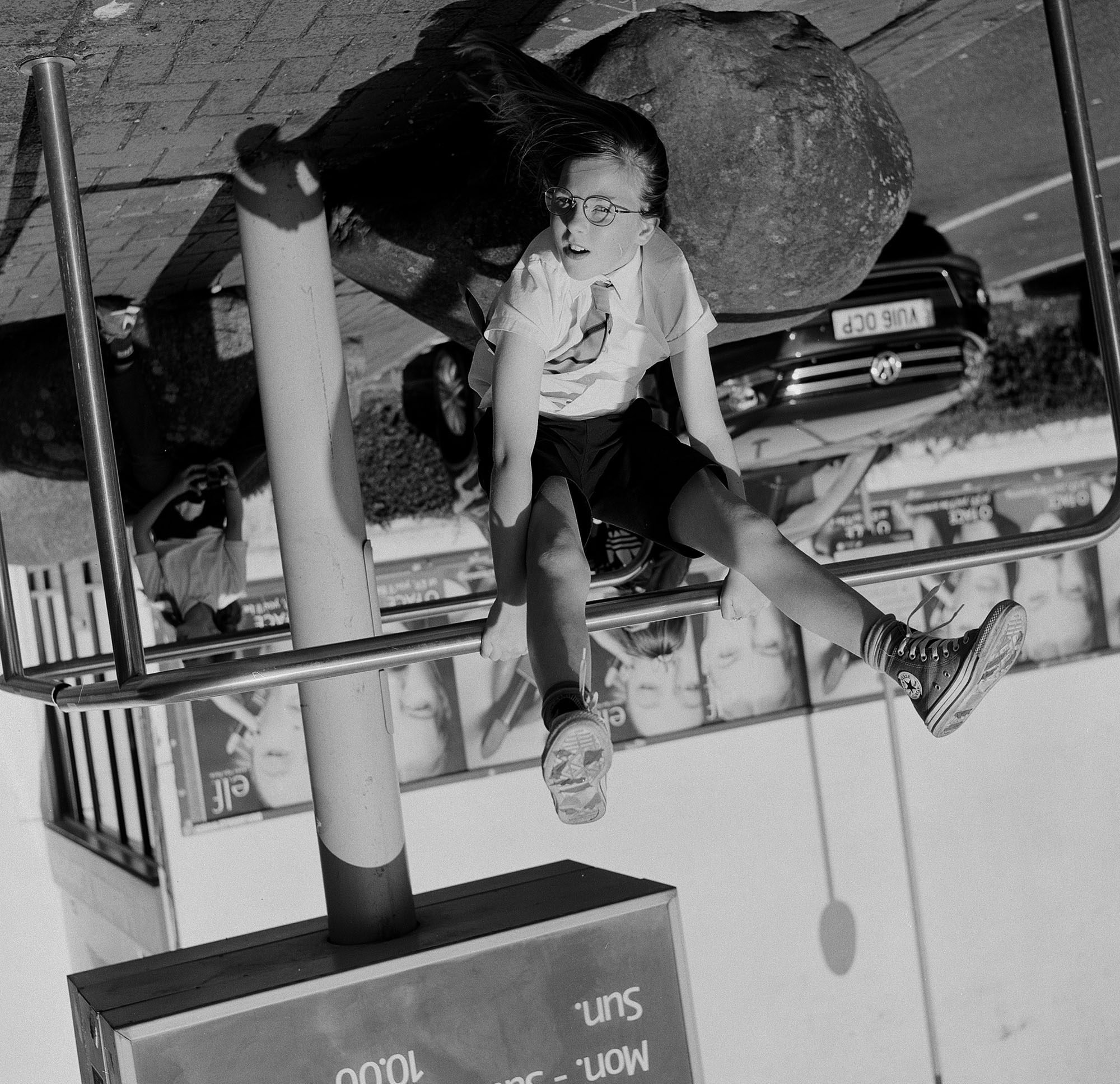 School child in uniform hangs on a metal railing in a retail car park. The image is displayed upside down so that she appears upright while another young person watching from behind appears upside down.