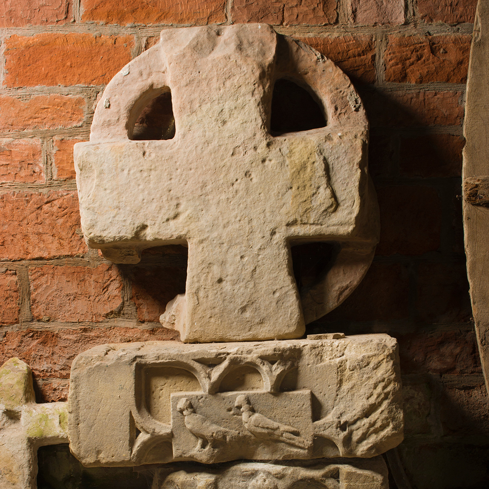 A large sandstone cross rests against a red brick wall, on top of a block carved with birds.
