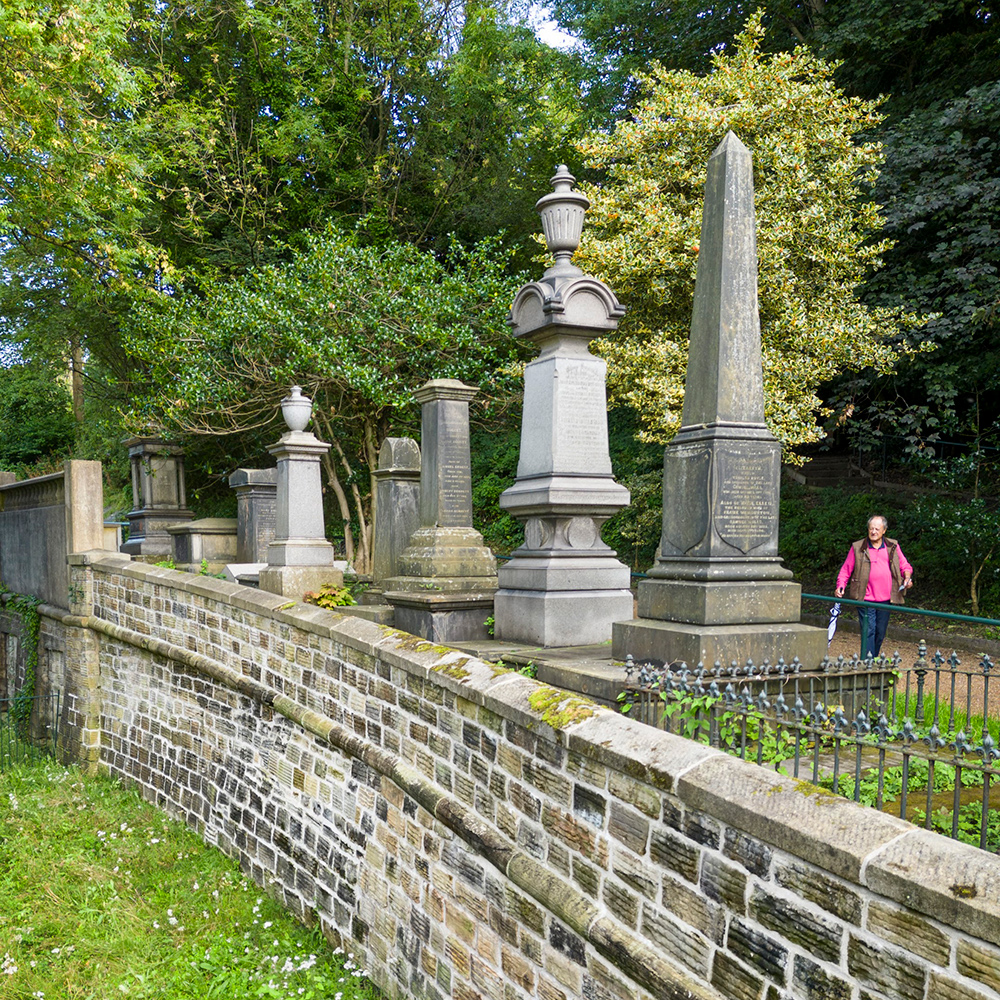 View of the exterior of a cemetery wall, with monuments and trees beyond. 
