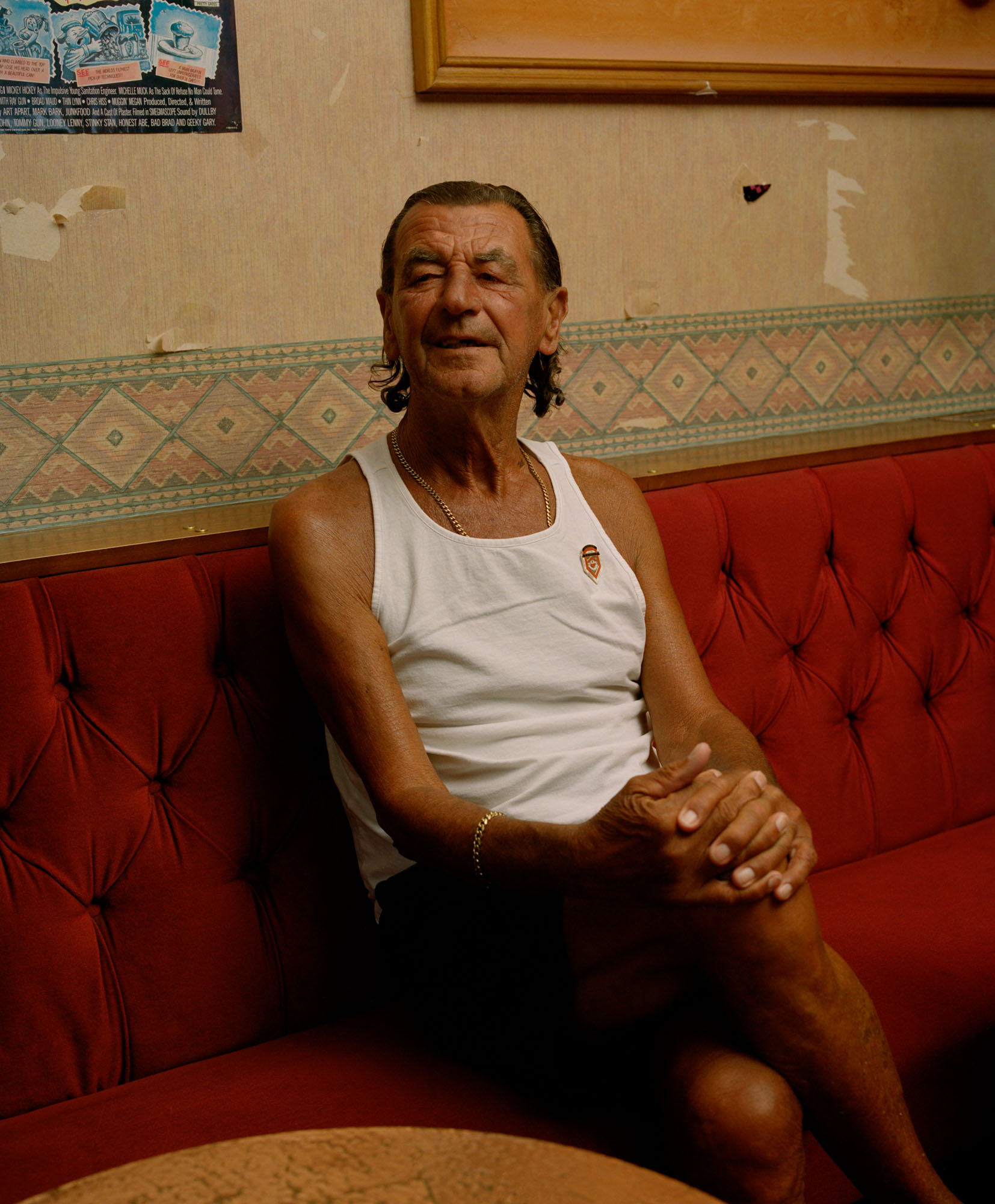 Person wearing a white tank top gold chain bracelet and necklace sits on red booth seating at a men's working club. There is patterned wallpaper behind them.