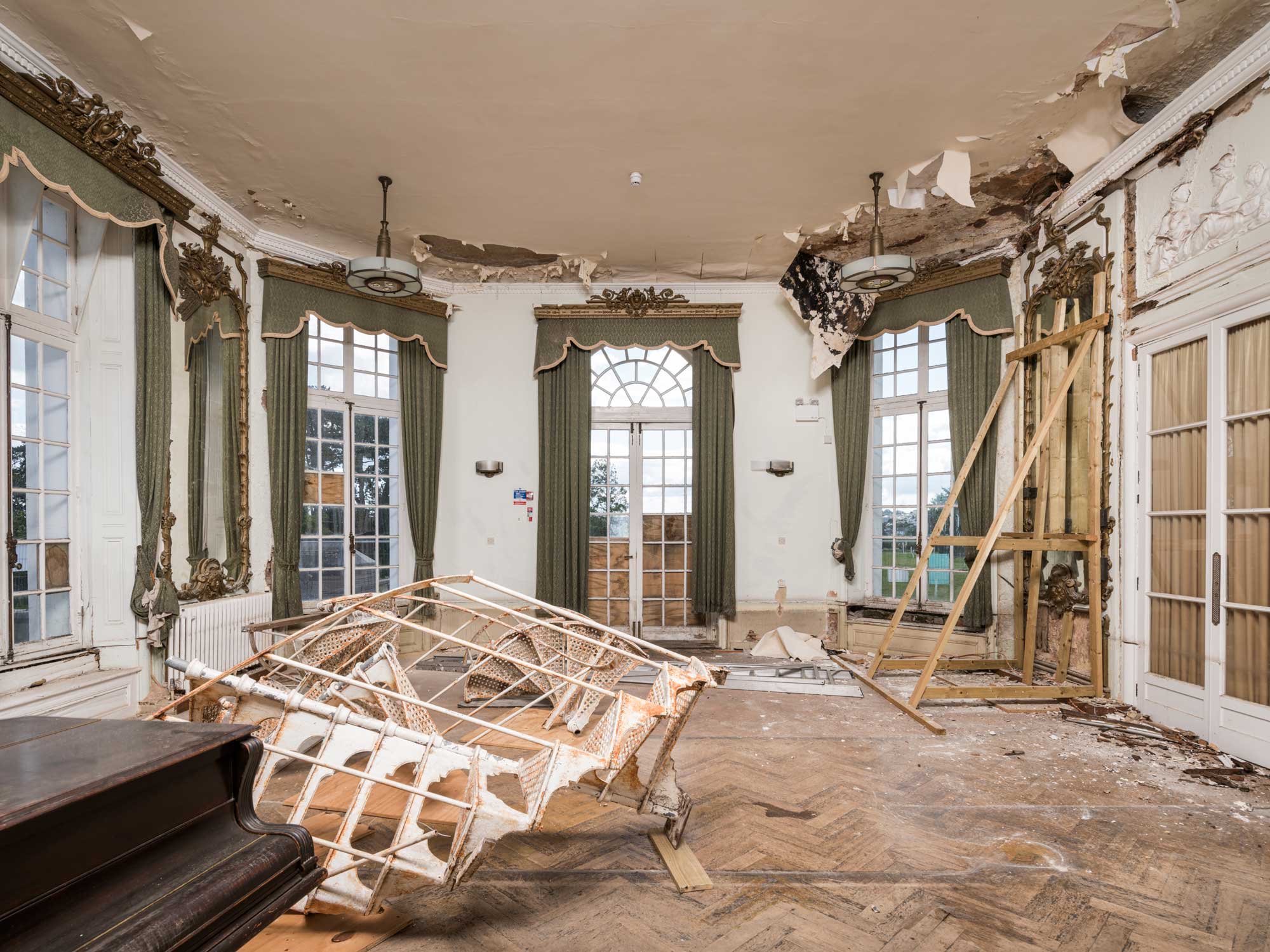 The derelict interior of a formal mansion room, with debris on the parquet flooring, a piano, and holes in the plasterwork. 