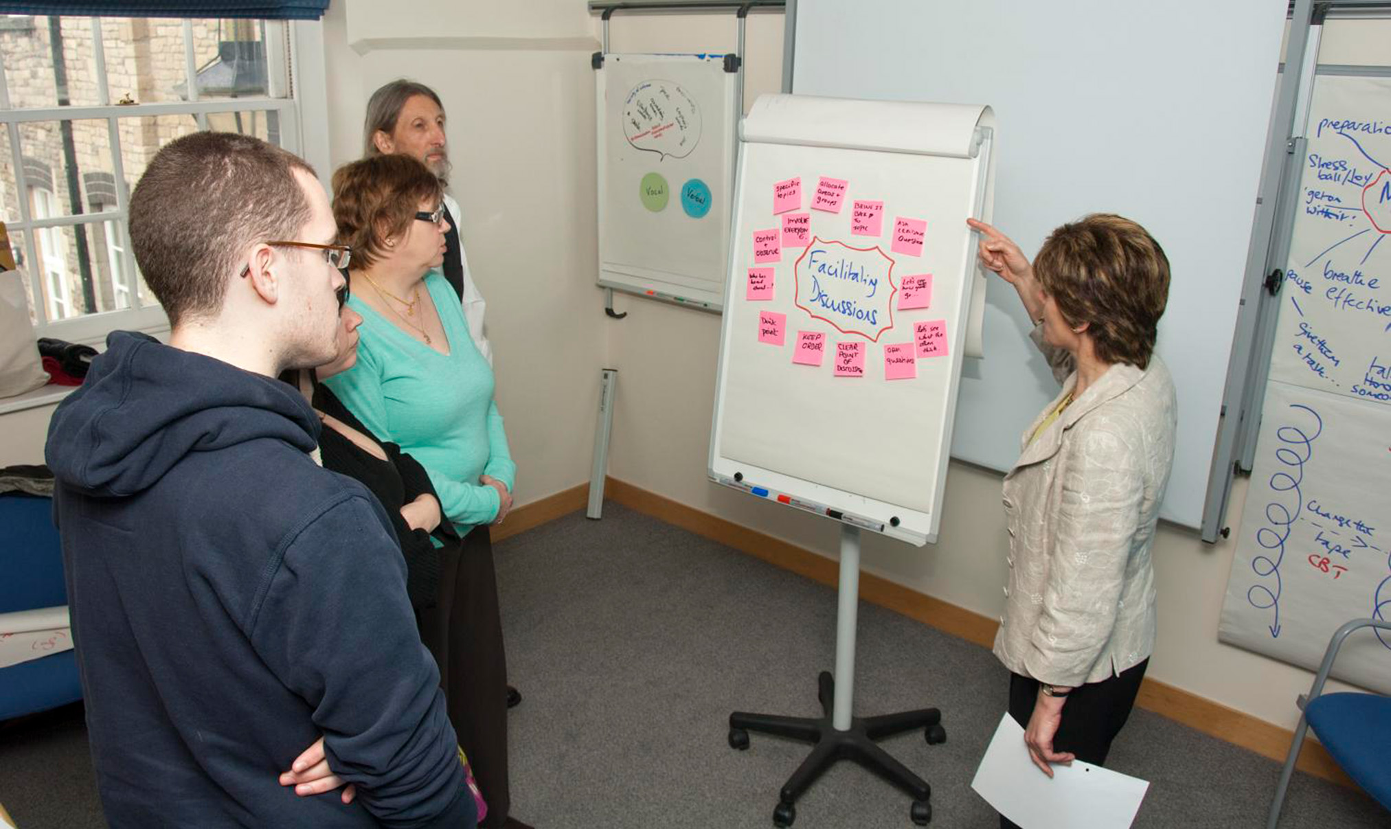 Four people gather around a paper flip chart on a stand and one person points to pink post it notes on the paper around the title, Facilitating Discussions