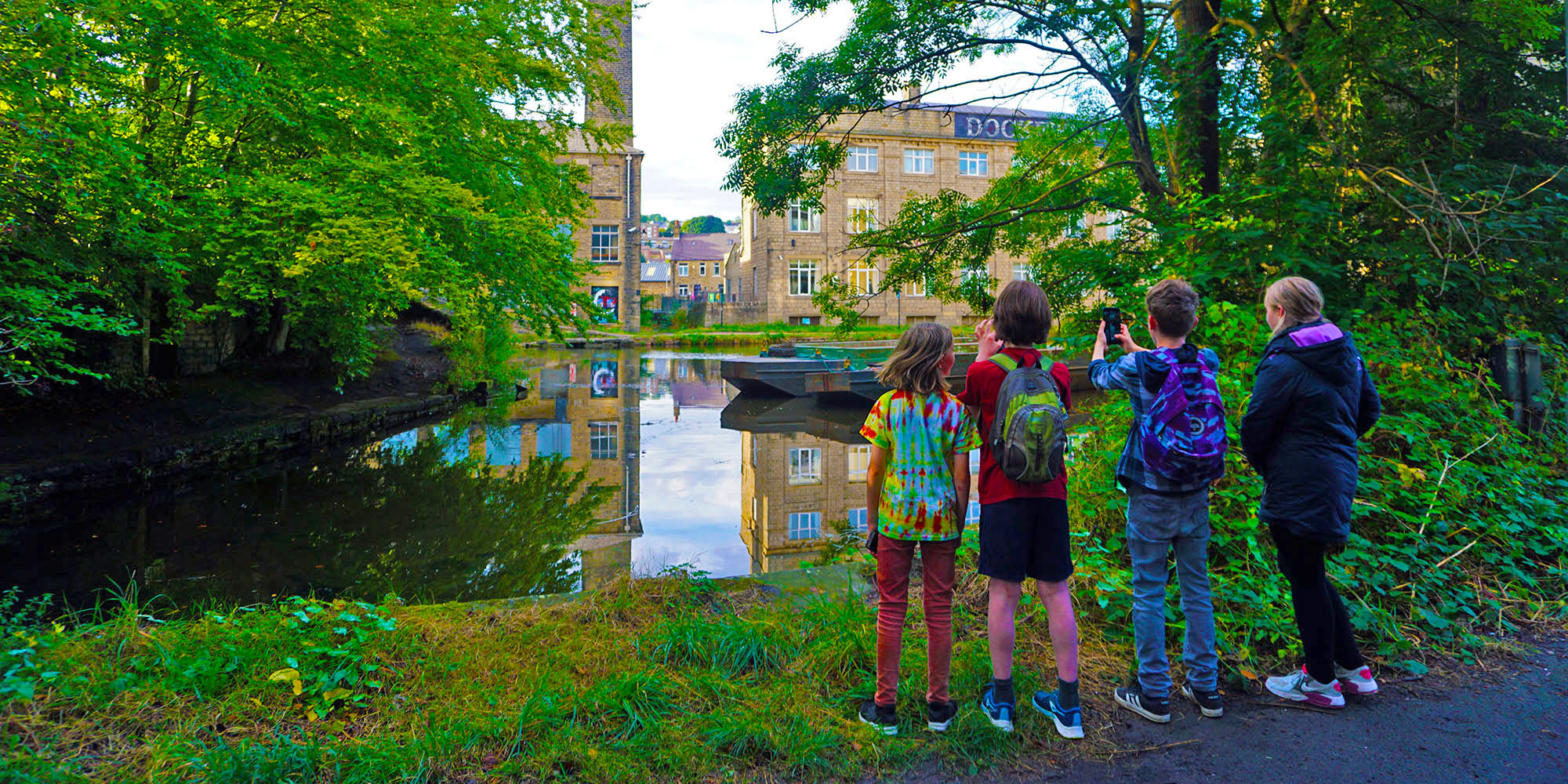 Three children stand on a canal bank, photographing the canal and industrial landscape through overhanging trees.