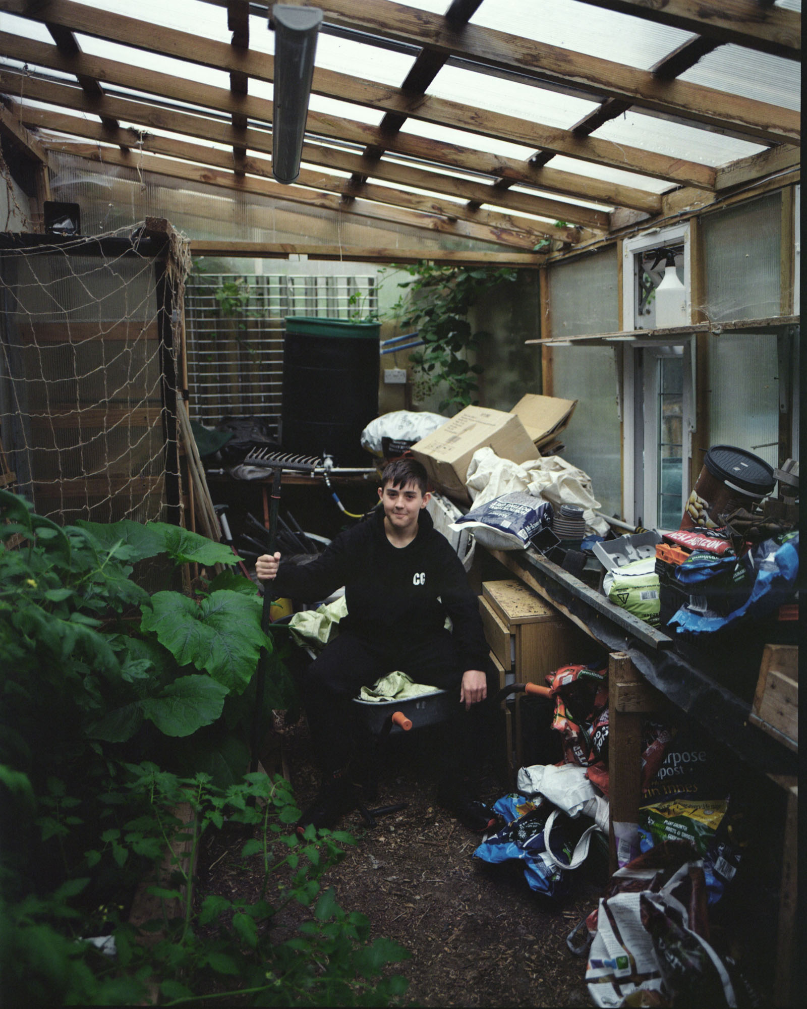 Boy sits in a greenhouse surrounded by plants, boxes and gardening tools.