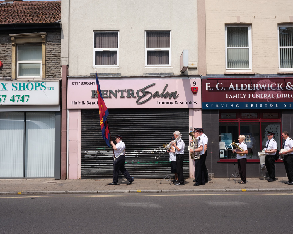 People in Salvation Army uniforms carry a flag and play brass band instruments down a high street.