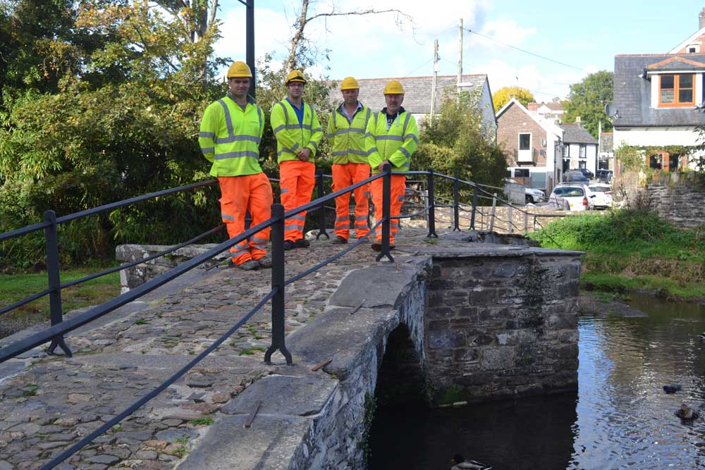 A photograph of four people on an ancient stone bridge across a river. They are wearing hi-vis clothes and hard hats.