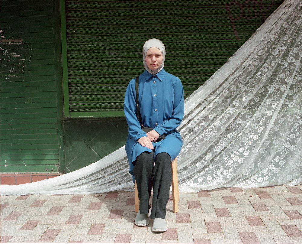 A woman sits for a photo in front of a shuttered shop front with white net curtain fabric draped behind her.
