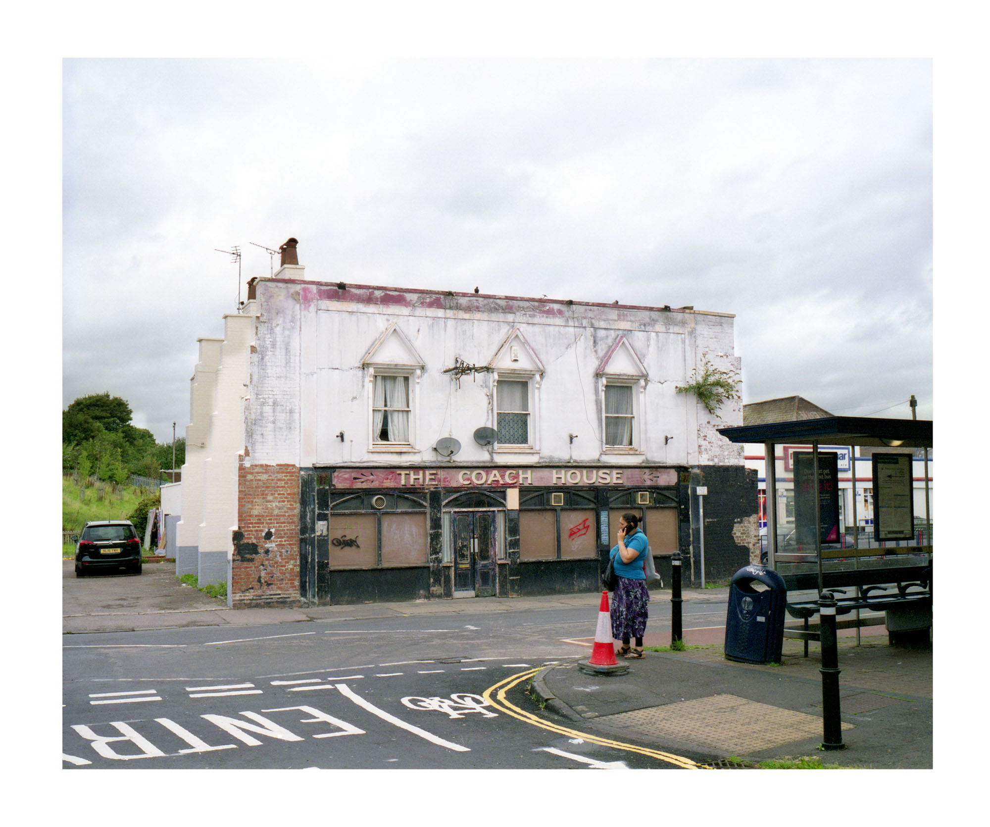 An old boarded up pub called The Coach House viewed across a road. In the foreground a woman stands next to a traffic cone talking on her phone.