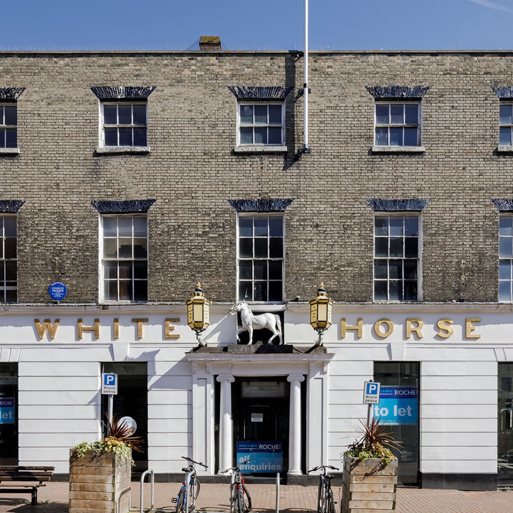 Three storey 19th century building, with white exterior at the bottom and stone exterior on the top two levels. The entrance has Great White Horse Hotel in gold letter, lanterns and a sculptured white horse above a main entrance with pillars.