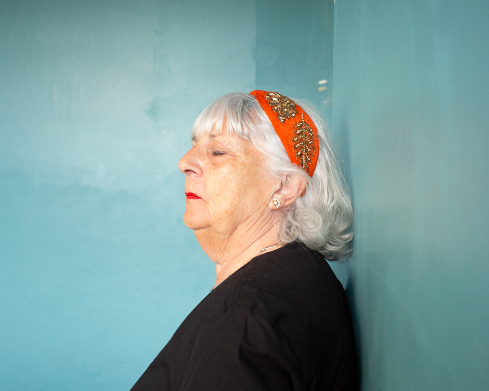 Head and shoulders photo portrait of a woman in profile. She wears an orange headband and red lipstick, and she has her eyes shut.