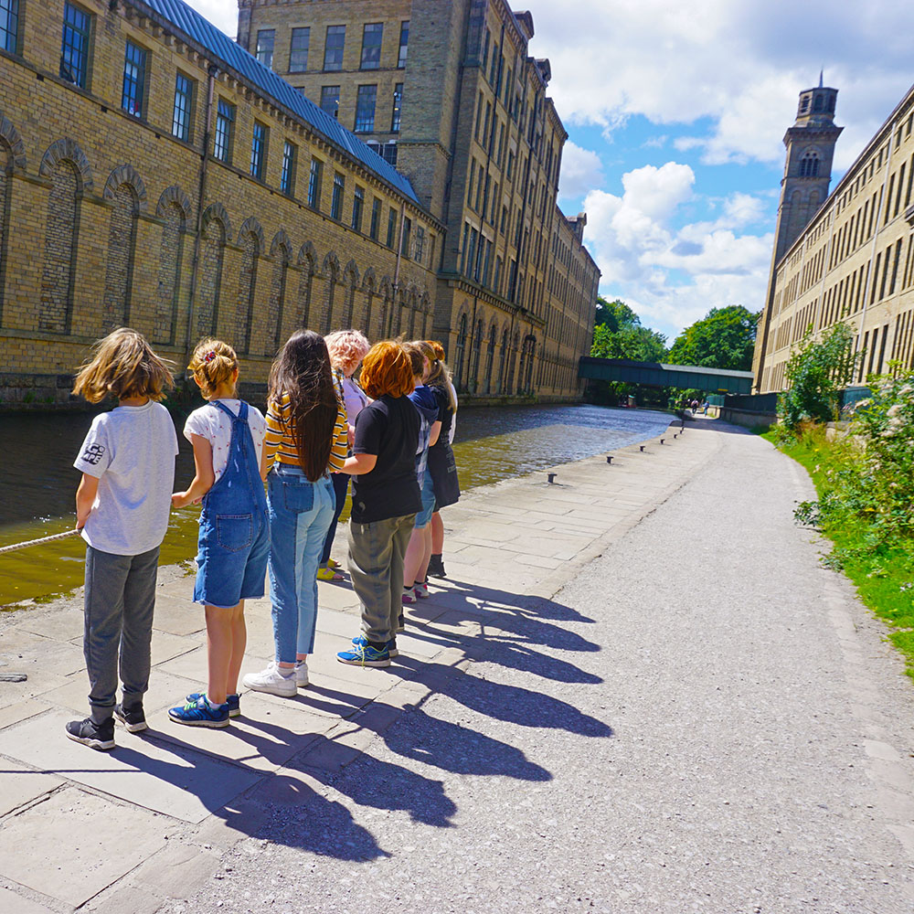A group of children and one adult hold a rope on a canal towpath in an industrial landscape.