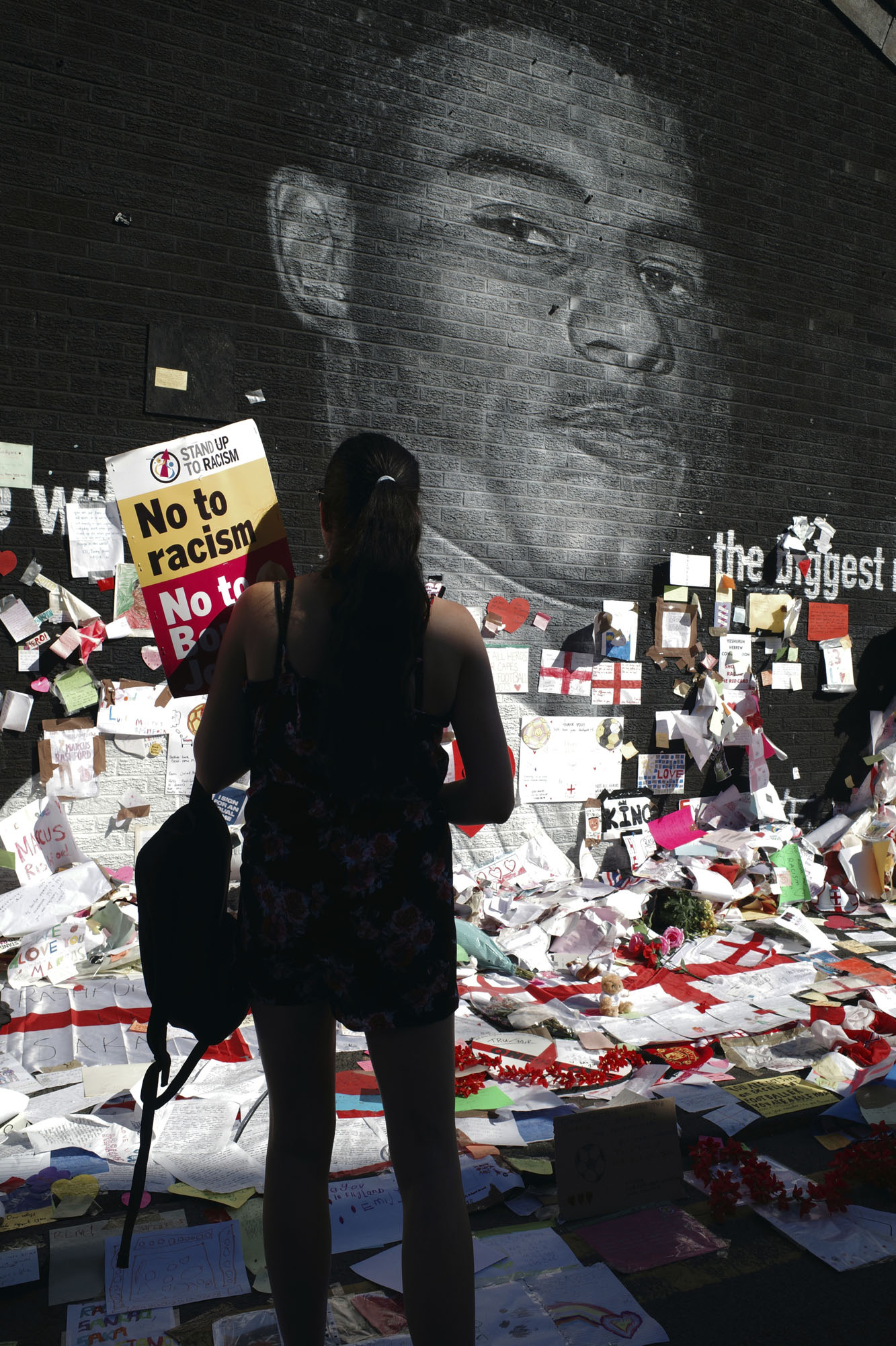 A woman standing in front of the Marcus Rashford mural in Withington, with posters, flowers and flags on the ground and wall.