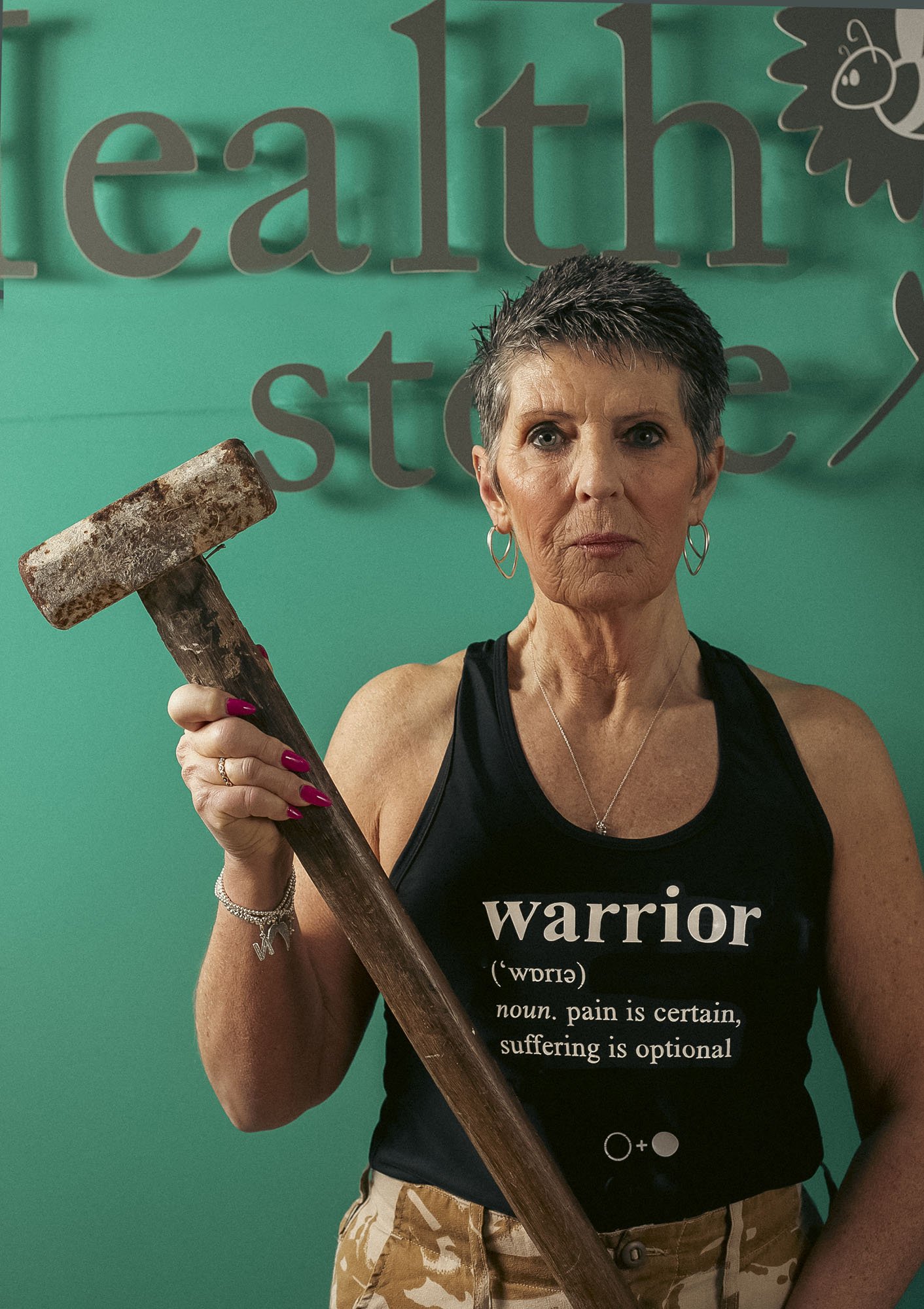 Portrait of a woman, holding a sledgehammer and wearing a vest with logo ‘Warrior, noun, pain is certain, suffering is optional’. 