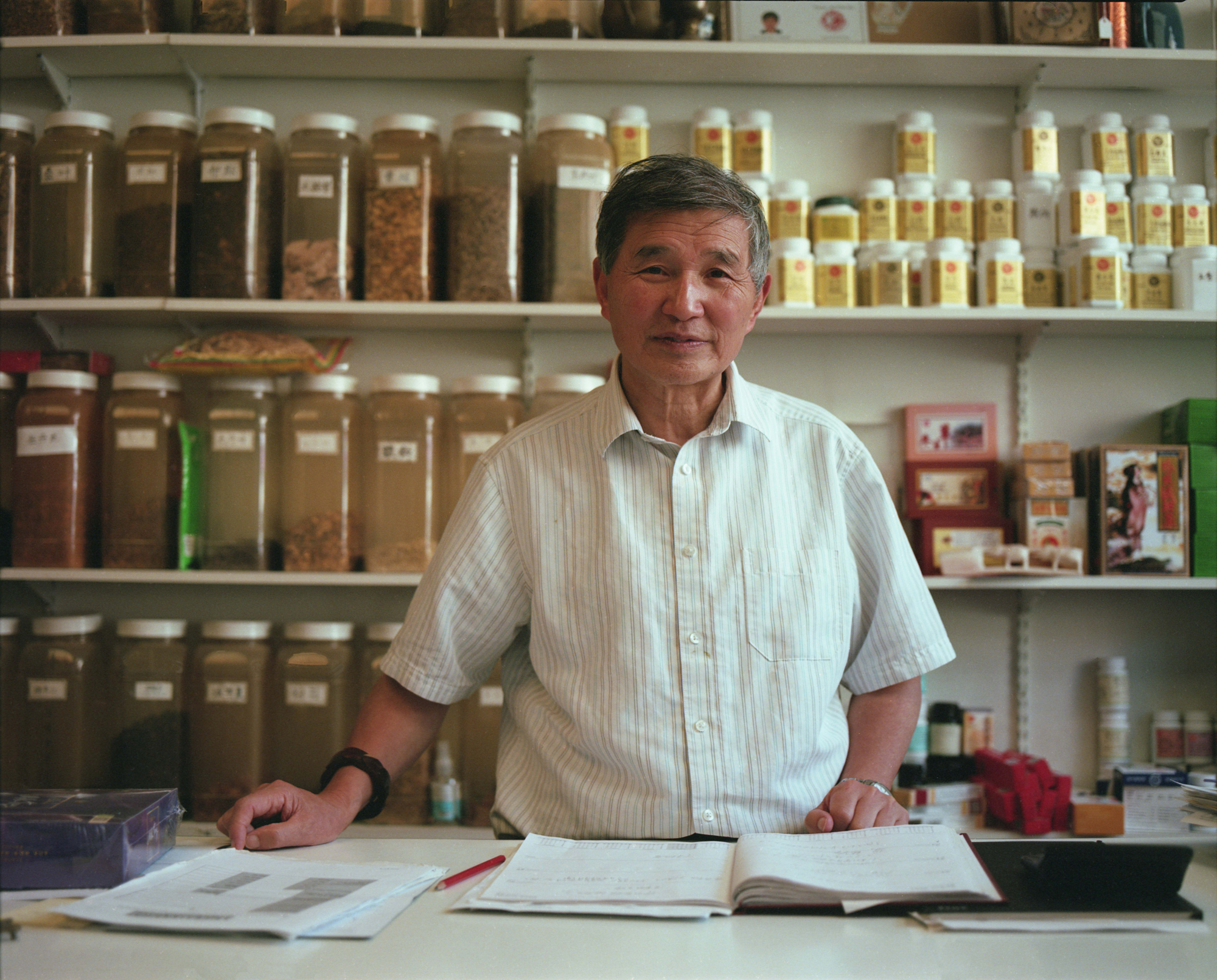 A man standing behind a counter. In front of him, on the counter is a ledger and papers and behind him 3 shelves are stacked with herbal products labelled in Chinese. 