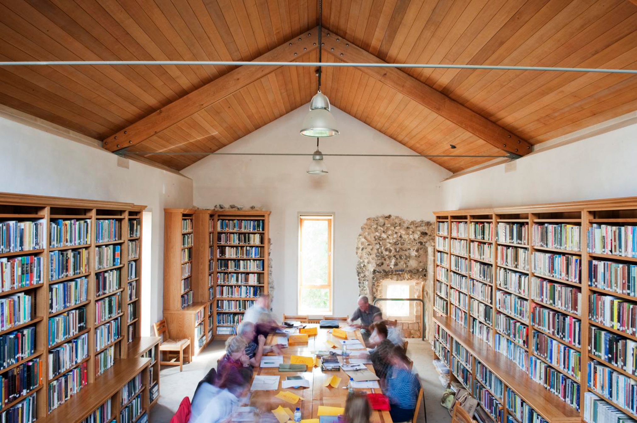 Bookshelves line the walls of a library with a wood panelled ceiling people work at a long table in the centre of the room. The people in the image are blurred by a long exposure and are unidentifiable.