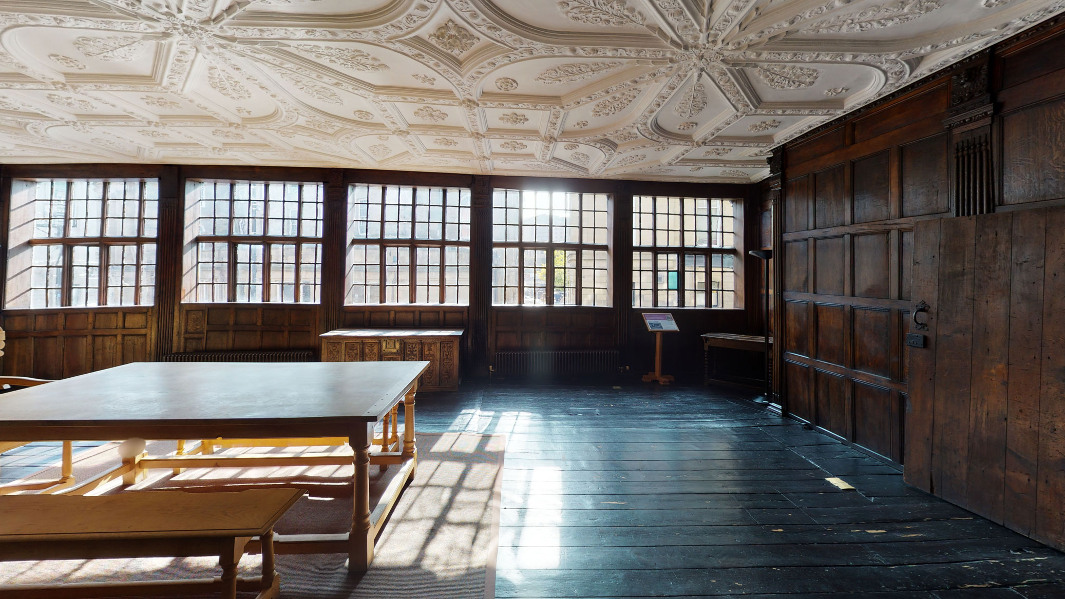A historic interior with wooden panelling and a stuccoed ceiling, illuminated by a range of historic windows.
