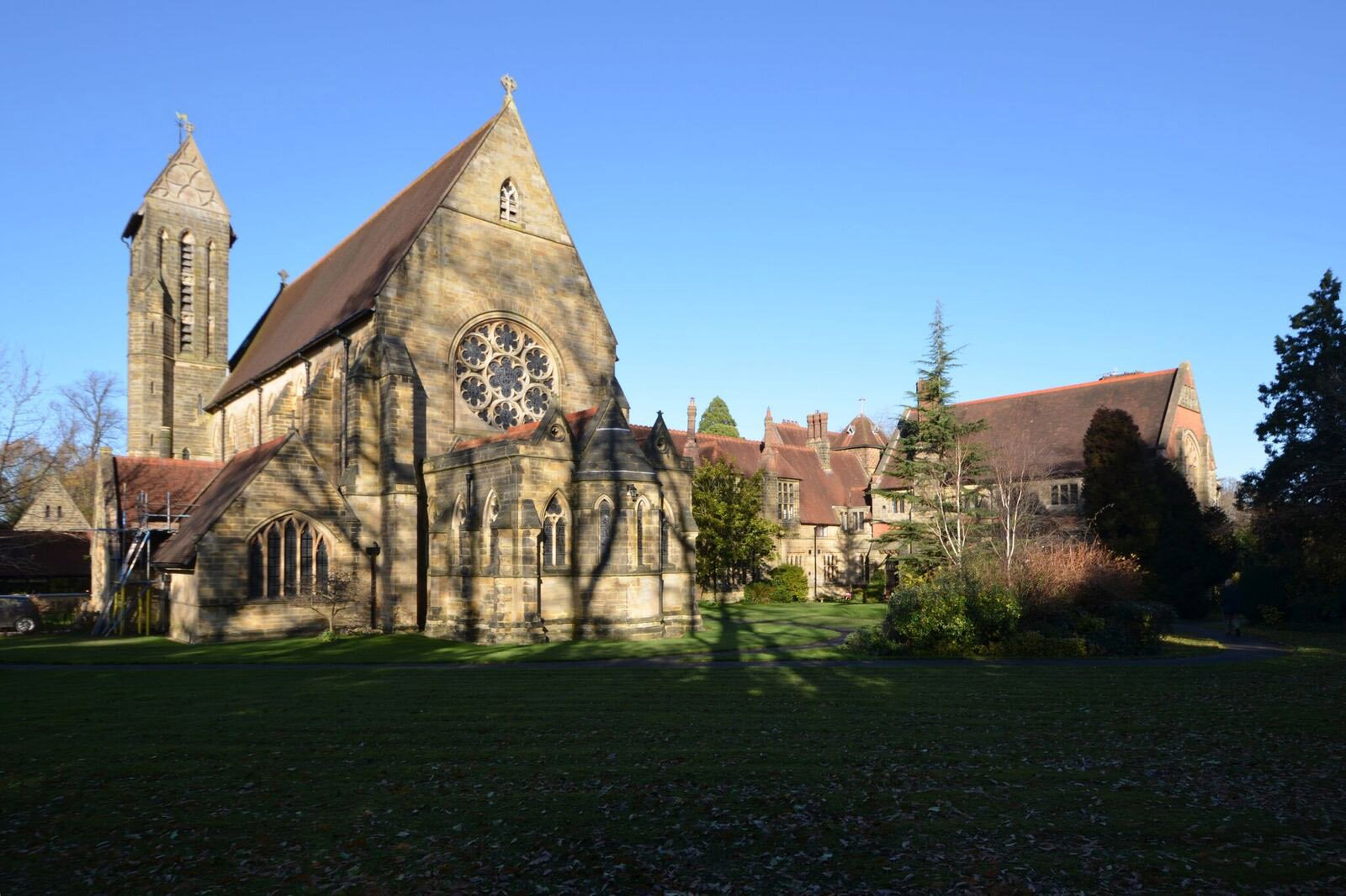 Group of stone buildings, including a church, with shadows cast on the lawn in the foreground and a clear blue sky above.