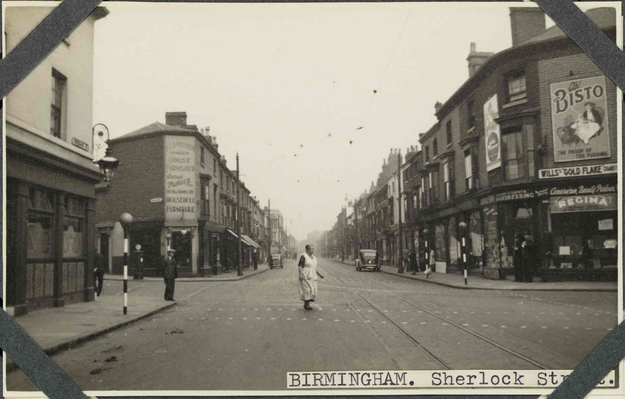 Black and white photograph of an urban street scene featuring a woman in an apron using a pedestrian crossing. Three-storey buildings flank the road, most of which have ground floor shops. The upper storeys of some buildings show painted signs and advertisement boards. A label applied to the bottom-right of the photograph reads: 'Birmingham. Sherlock Street', part of which is obscured by one of four corner photograph album mounts. 