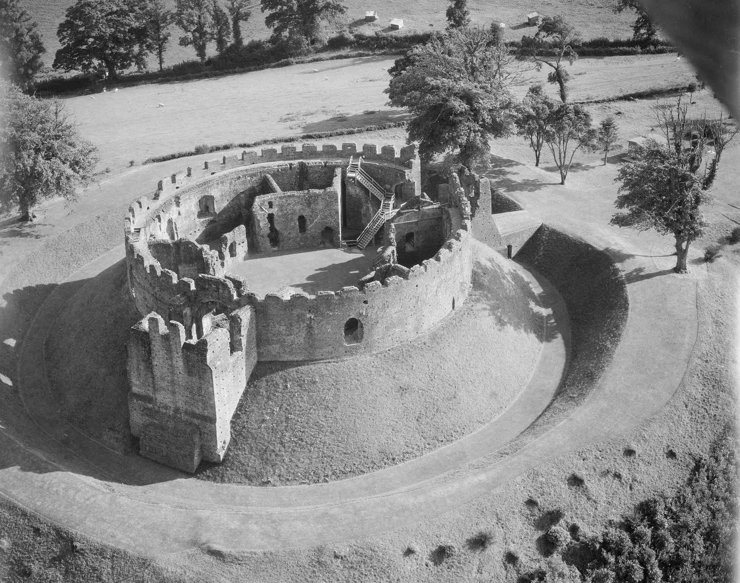Black and white oblique aerial photograph of the remains of a circular, crenelated, stone-walled castle, set on a grassy mound and surrounded by a ditch and bank. On the inside of the castle wall are rooms demarcated by the remains of walls, all of which are roofless. In the foreground, the remains of a square tower project from the curve of the castle wall.  
