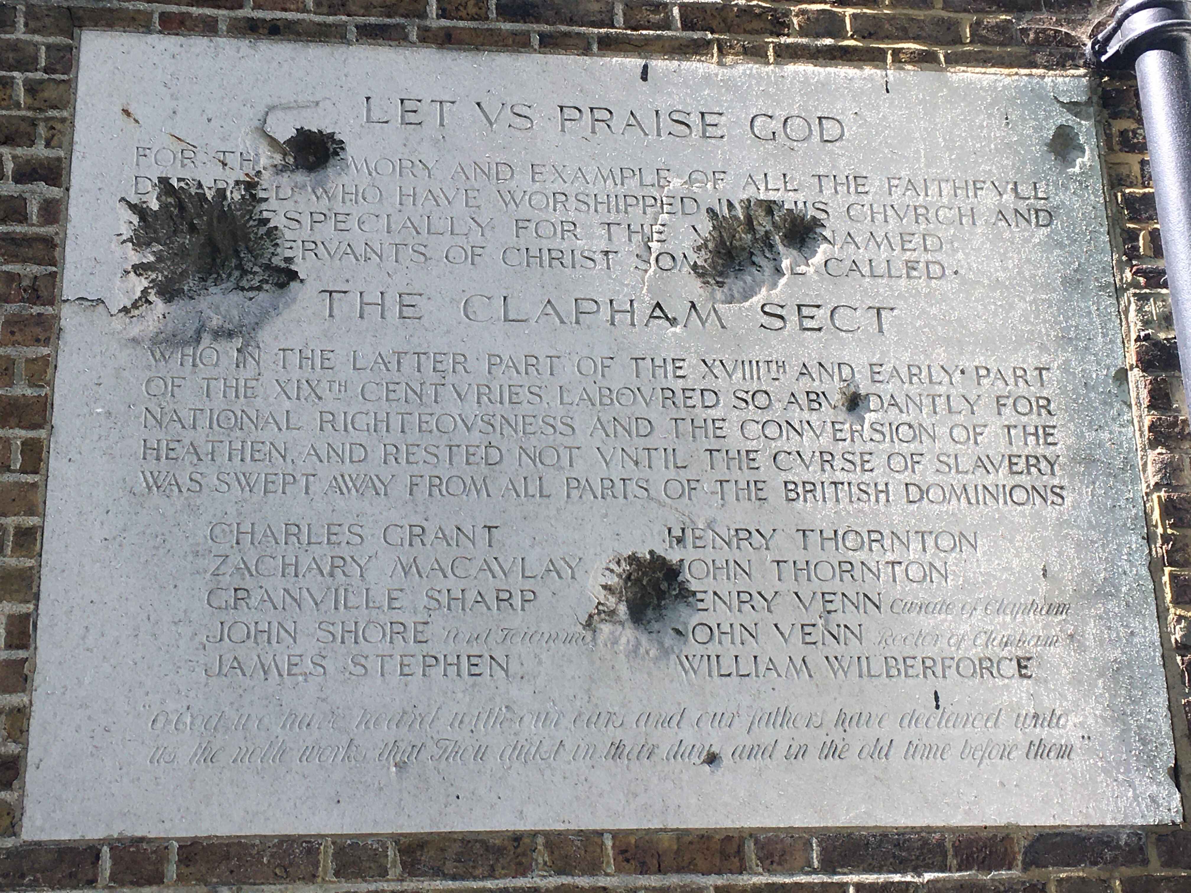 Stone plaque in brickwork. The plaque pitted with holes, partly obscuring the text. This includes the headings, Let us praise God and The Clapham Sect, with the names of the members ‘who…rested not until the curse of slavery was swept away’.