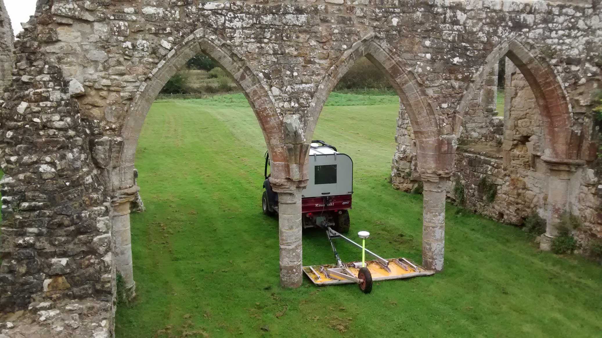 A vehicle towing geophysical survey equipment  passing through an arch within medieval ruins.