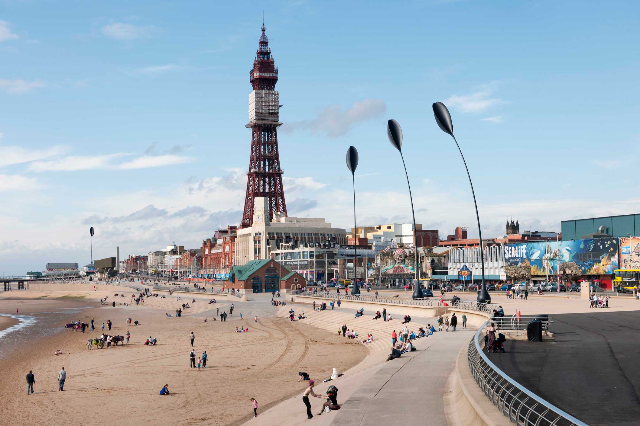 A 21st-century view of a beach, with seas defences, a promenade with tall sculptures in the shape of dune grass and Blackpool tower.
