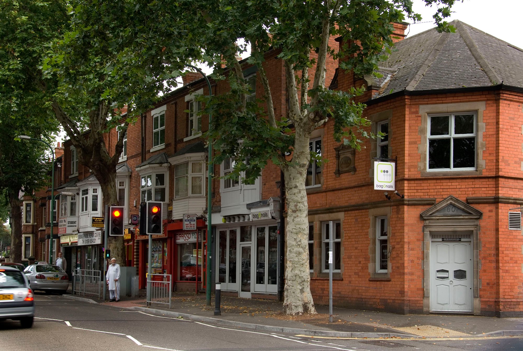 Road with a row of red brick buildings to the right. The pavement is lined by tall trees. A man wearing white is standing by the traffic lights.