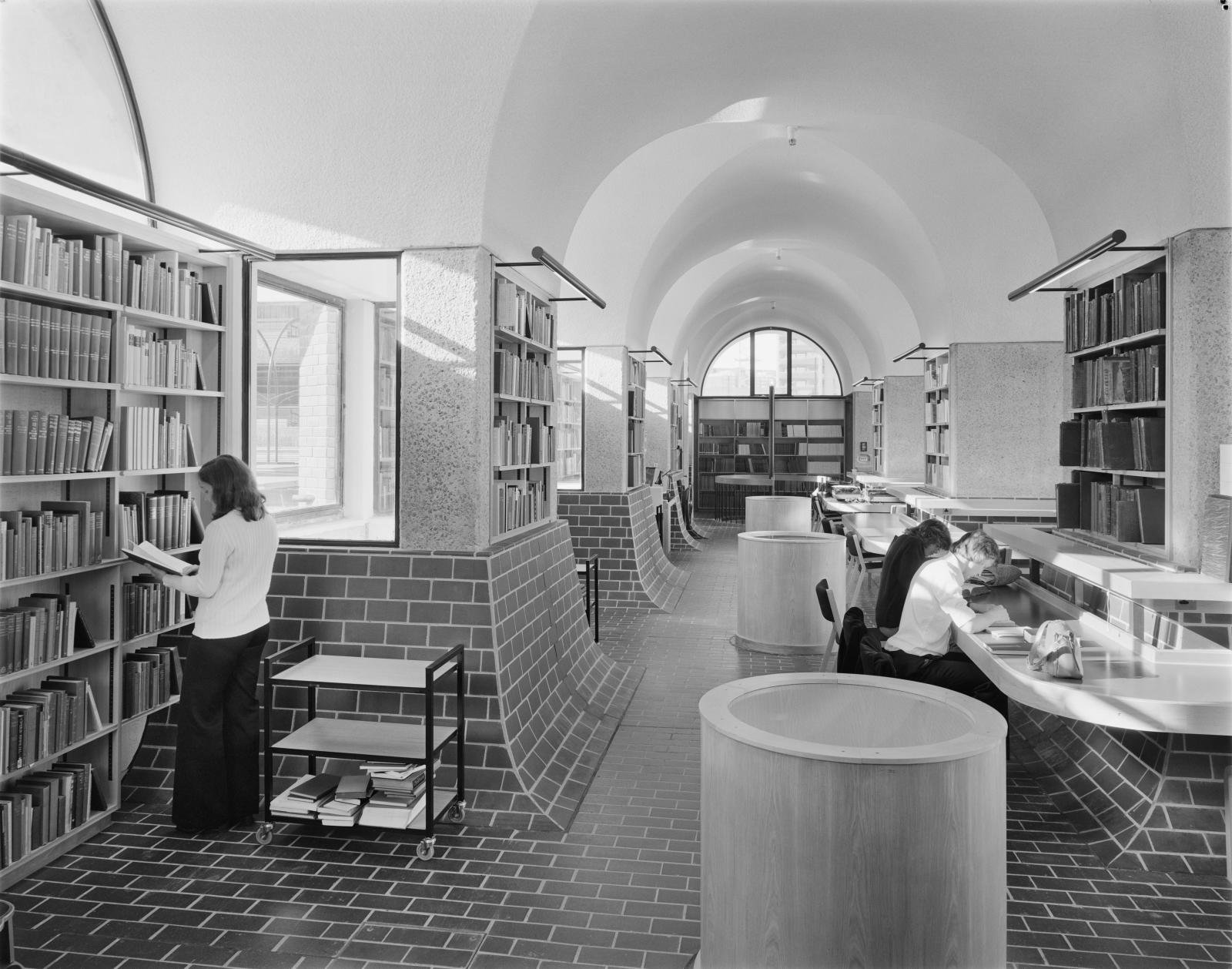 A black and white photo showing the interior of the Guildhall School of Music and Drama Library. The library’s floors and walls are partially covered with bricks, and three people are reading books in the area.