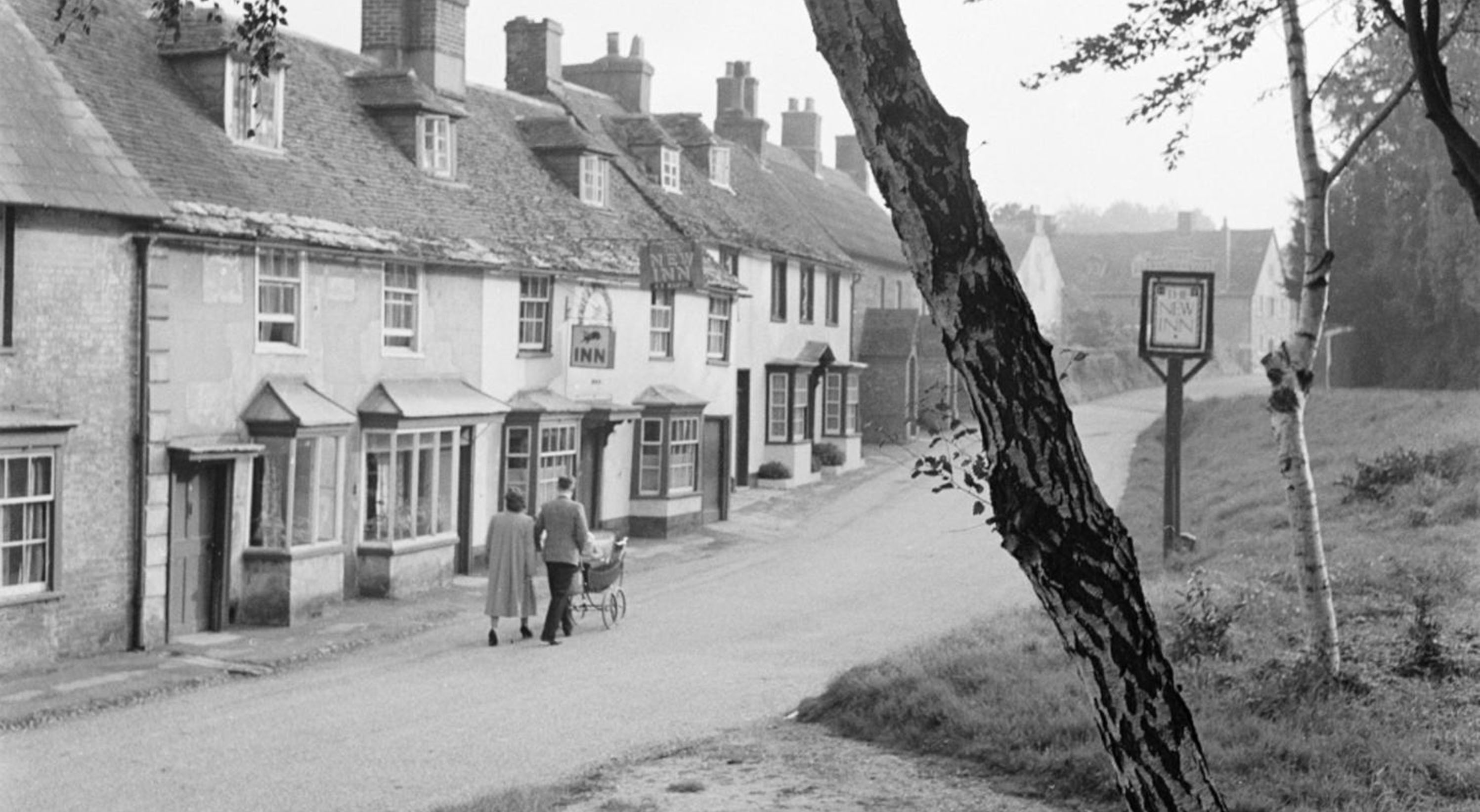 A man and a woman pushing a pram along Dorchester Hill and past the New Inn at Lower Bryanston.