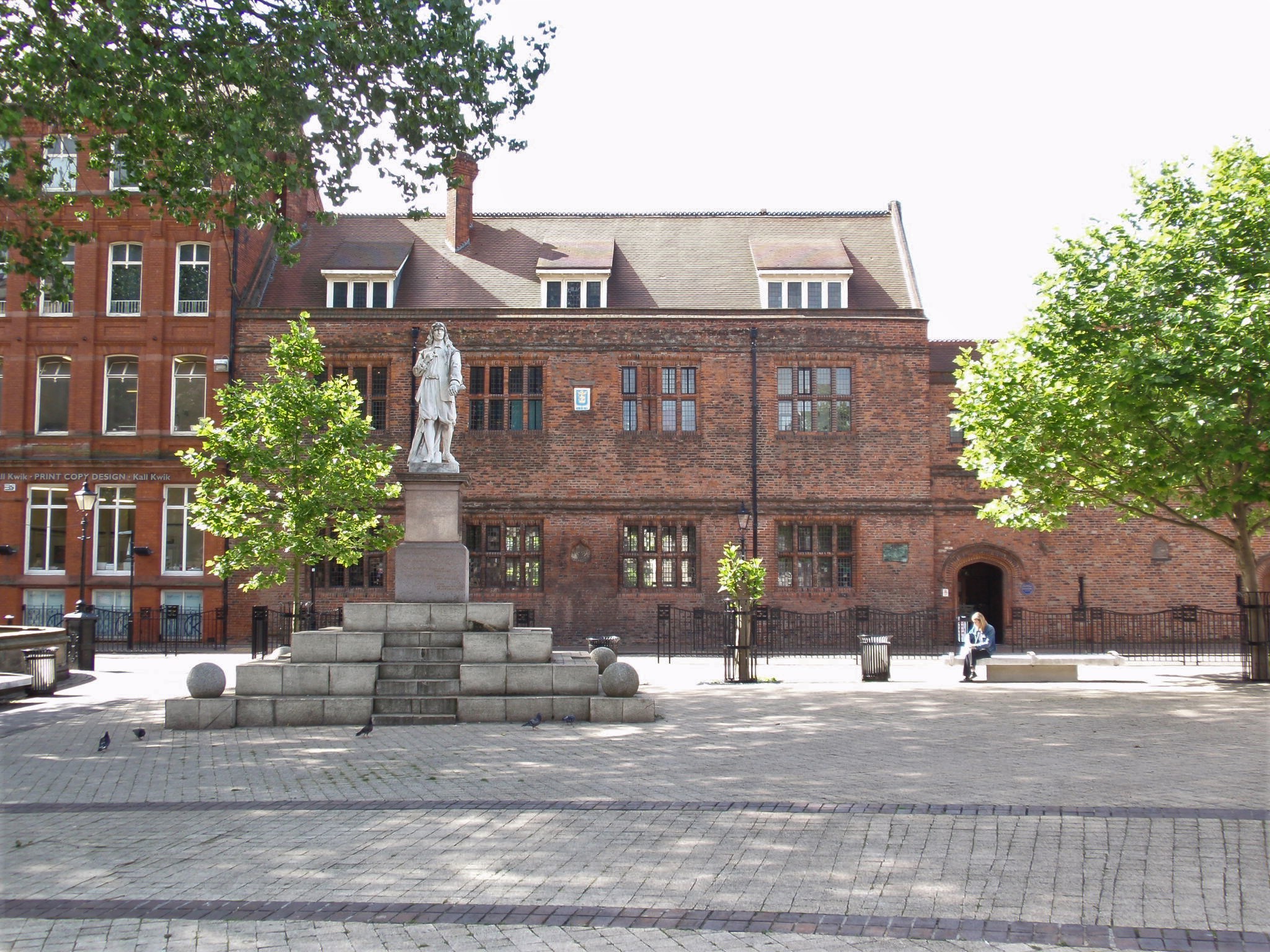 Statue in a paved square with a red brick building in the background and a couple of trees either side