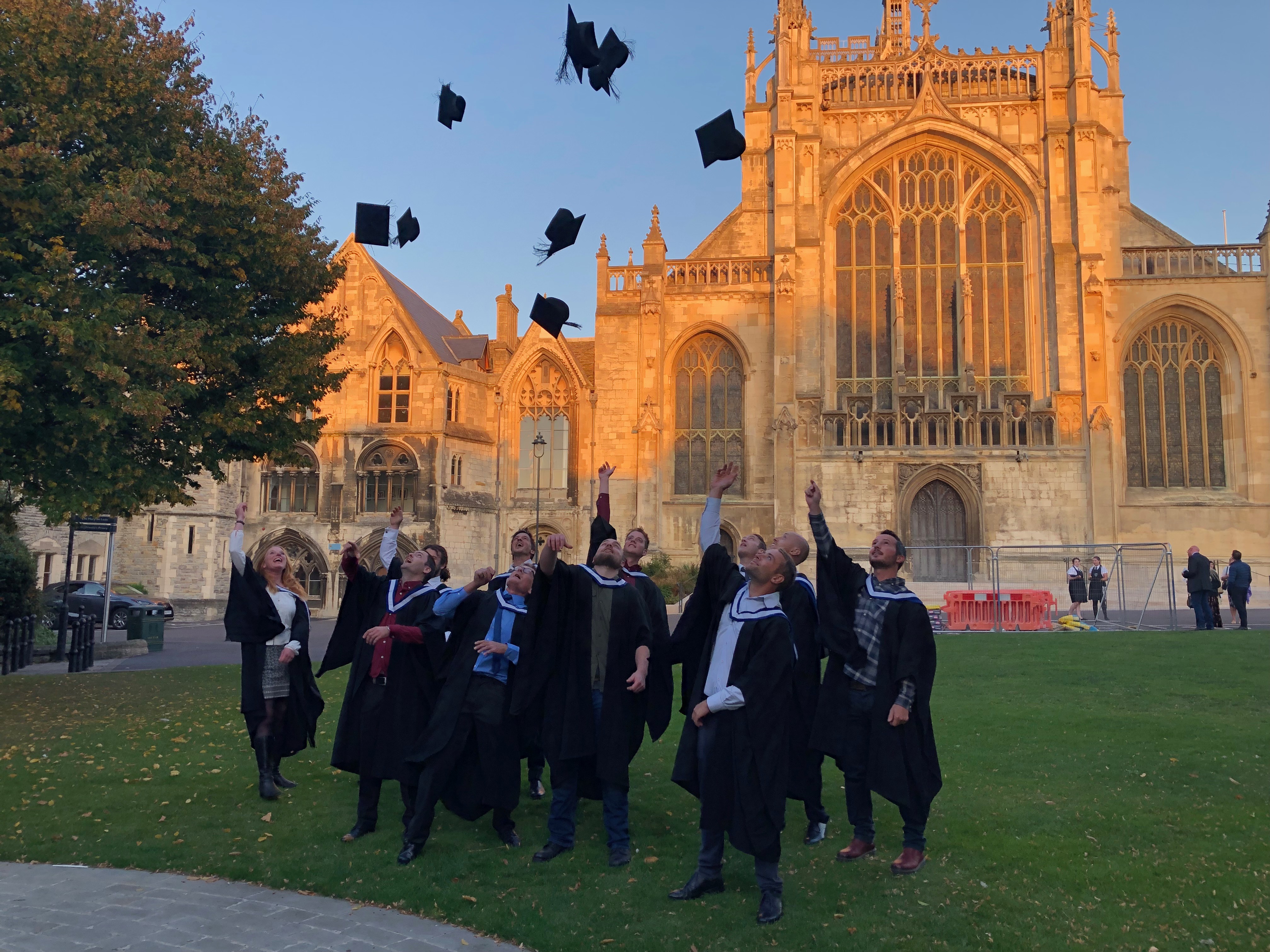 A group wearing graduation gowns throw their mortar boards in the air to celebrate. Behind them, light from the setting sun turns Gloucester Cathedral orange against a blue sky.