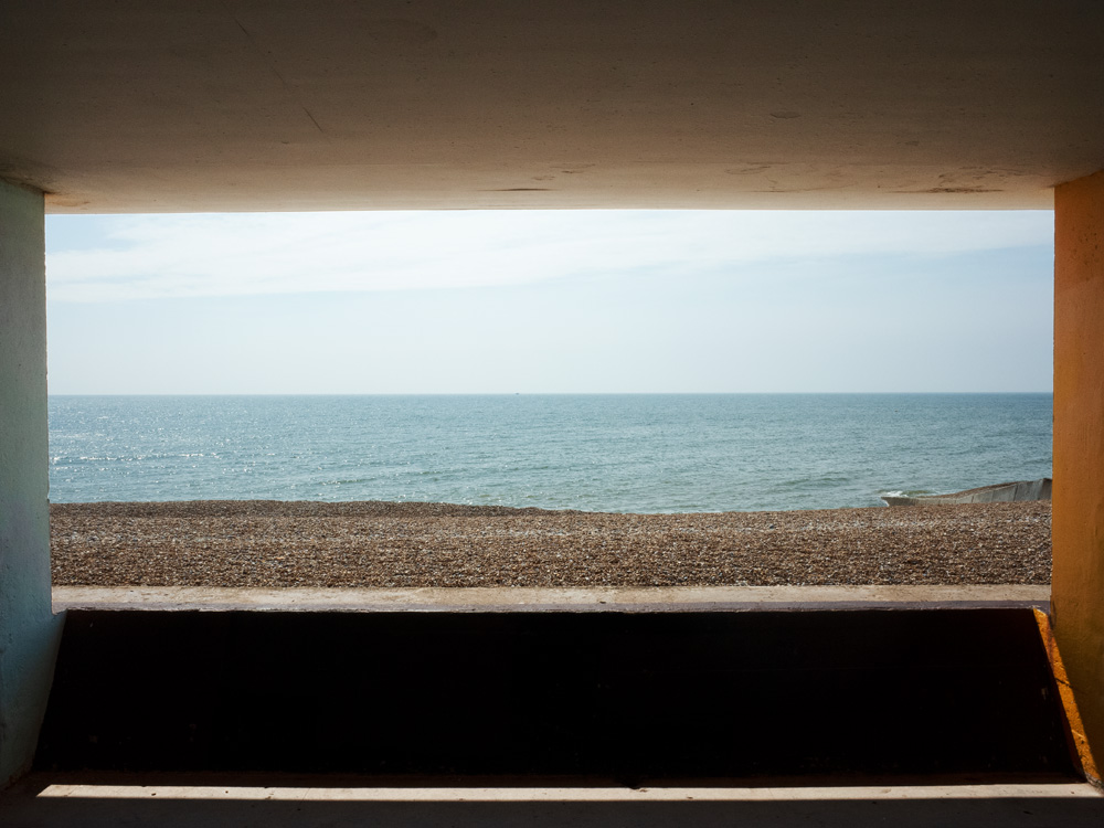 A view out to sea on a pebbly beach is framed by four concrete walls of a promenade.