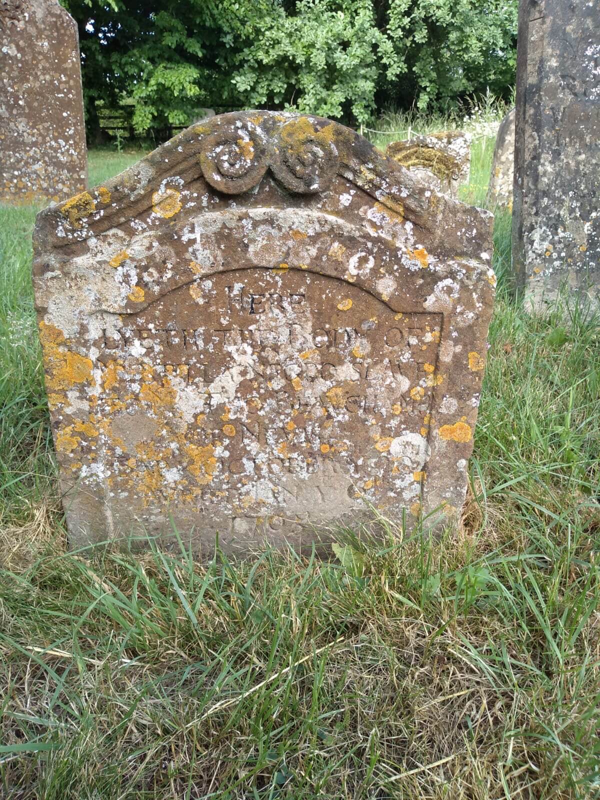 Headstone with scrolled detail to the top, the brown stone mottled with lichen. The indistinct inscription begins ‘Here lieth the body of Myrtilla negro slave’ and ends ‘baptised October 20th, buried January 6th 1705’.