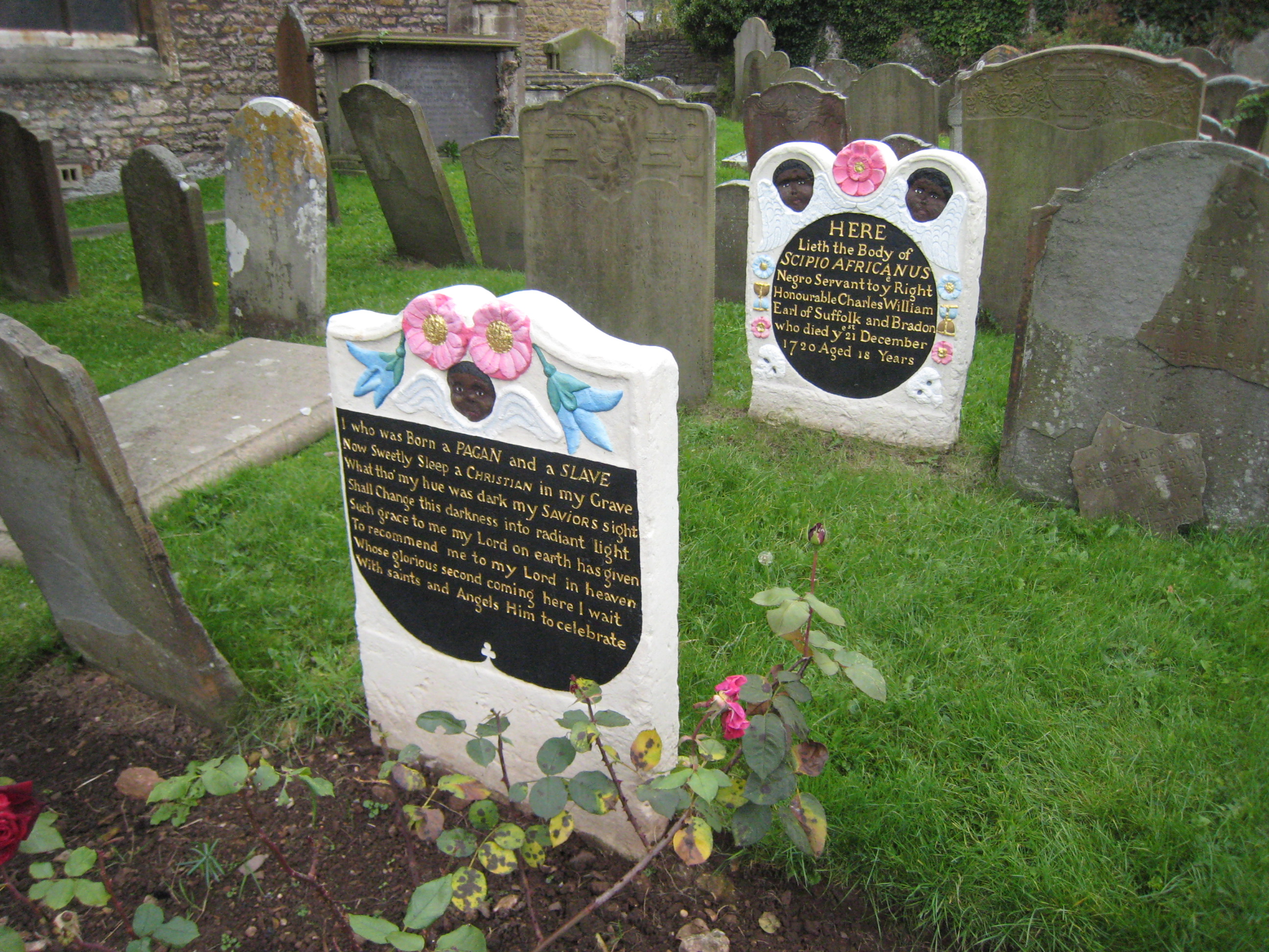 Headstone and footstone, with shaped tops. The freshly painted memorial stands out amongst unpainted stones. Carving includes winged Black cherubs, and flowers. The headstone carries the details of the subject’s life; the footstone carries a verse.