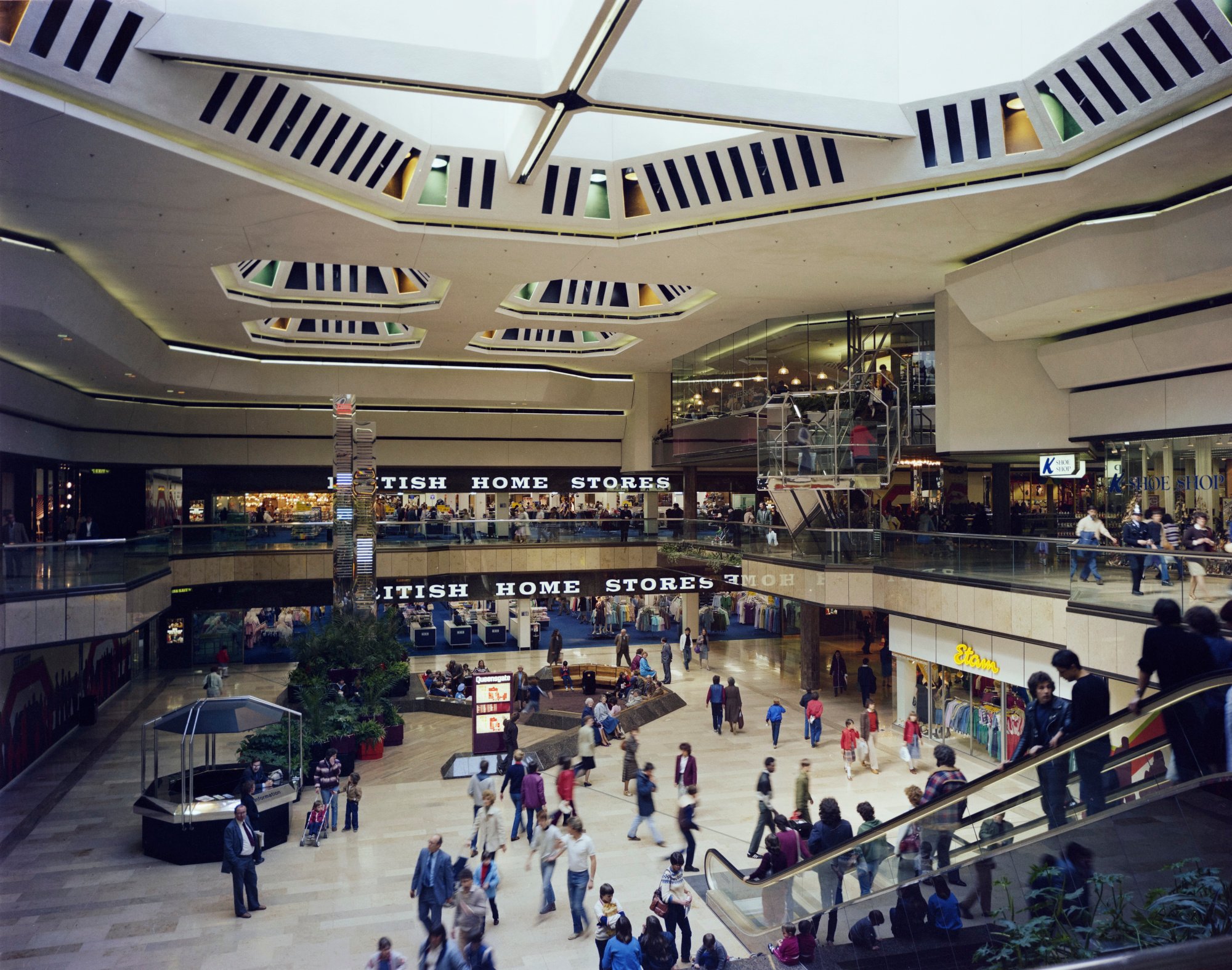 Colour photograph showing shoppers inside Queensgate shopping centre. The two levels of British Home Stores are in the background. On the right, shoppers are riding down an escalator into a large central square.