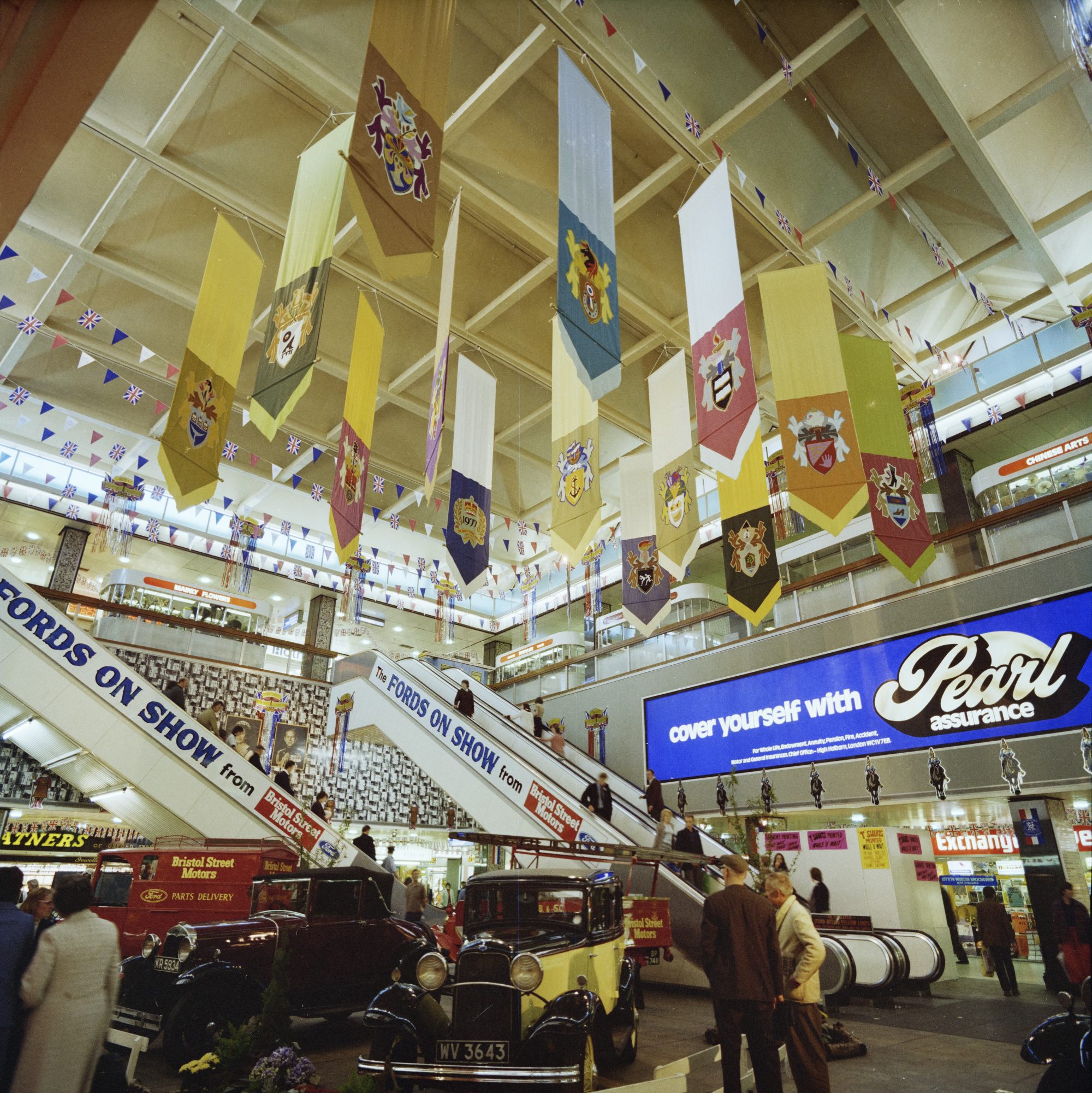 Colour photograph showing the Centre Court of the Bull Ring shopping centre. Vintage cars are on display as part of the ‘Fords on Show’ exhibition from Bristol Street Motors, and flags are up for the Queen’s Silver Jubilee.