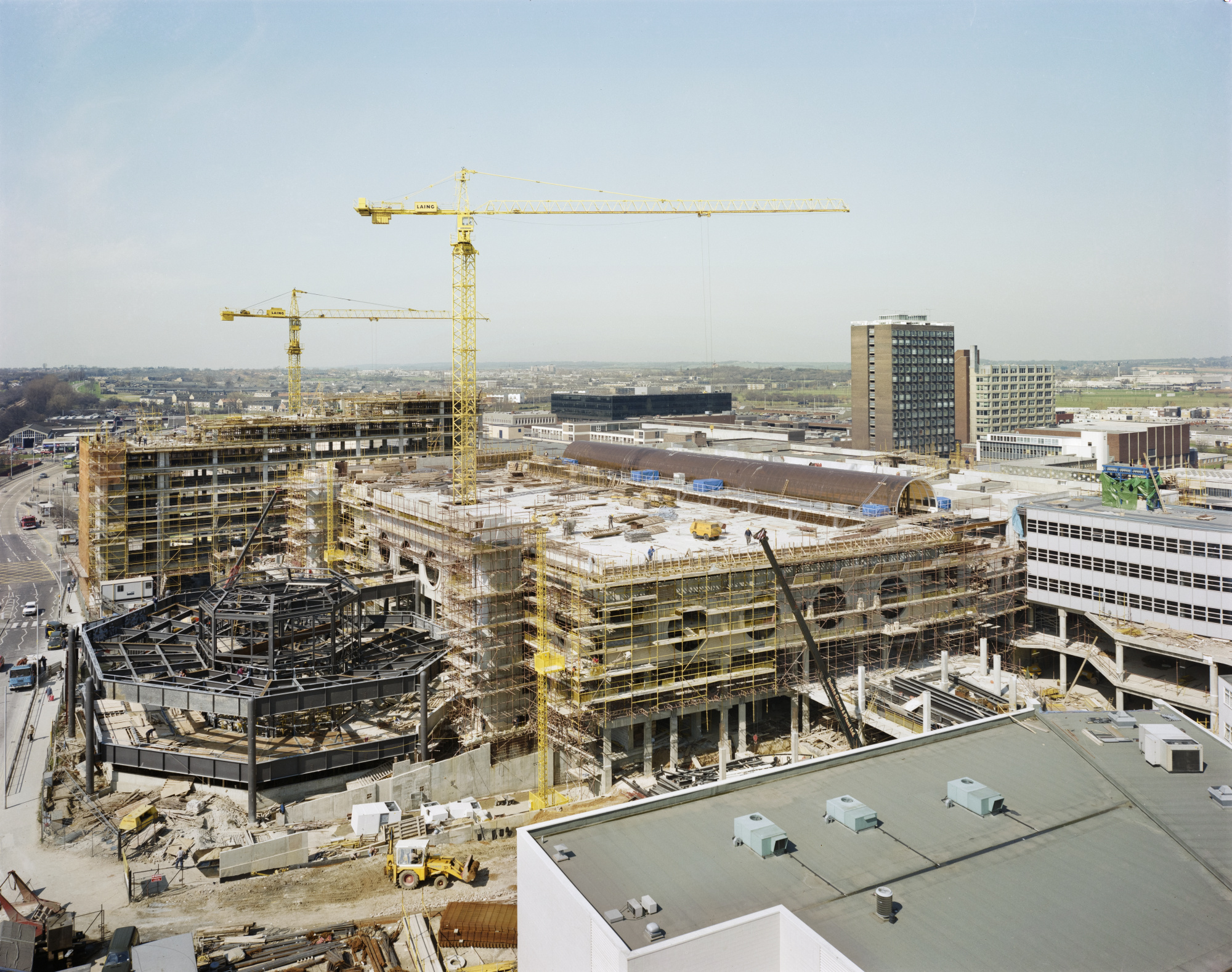 Colour photograph taken from the roof of a building showing the Eastgate Centre under construction. The partially constructed buildings are covered with scaffolding and two yellow cranes rise above the site.