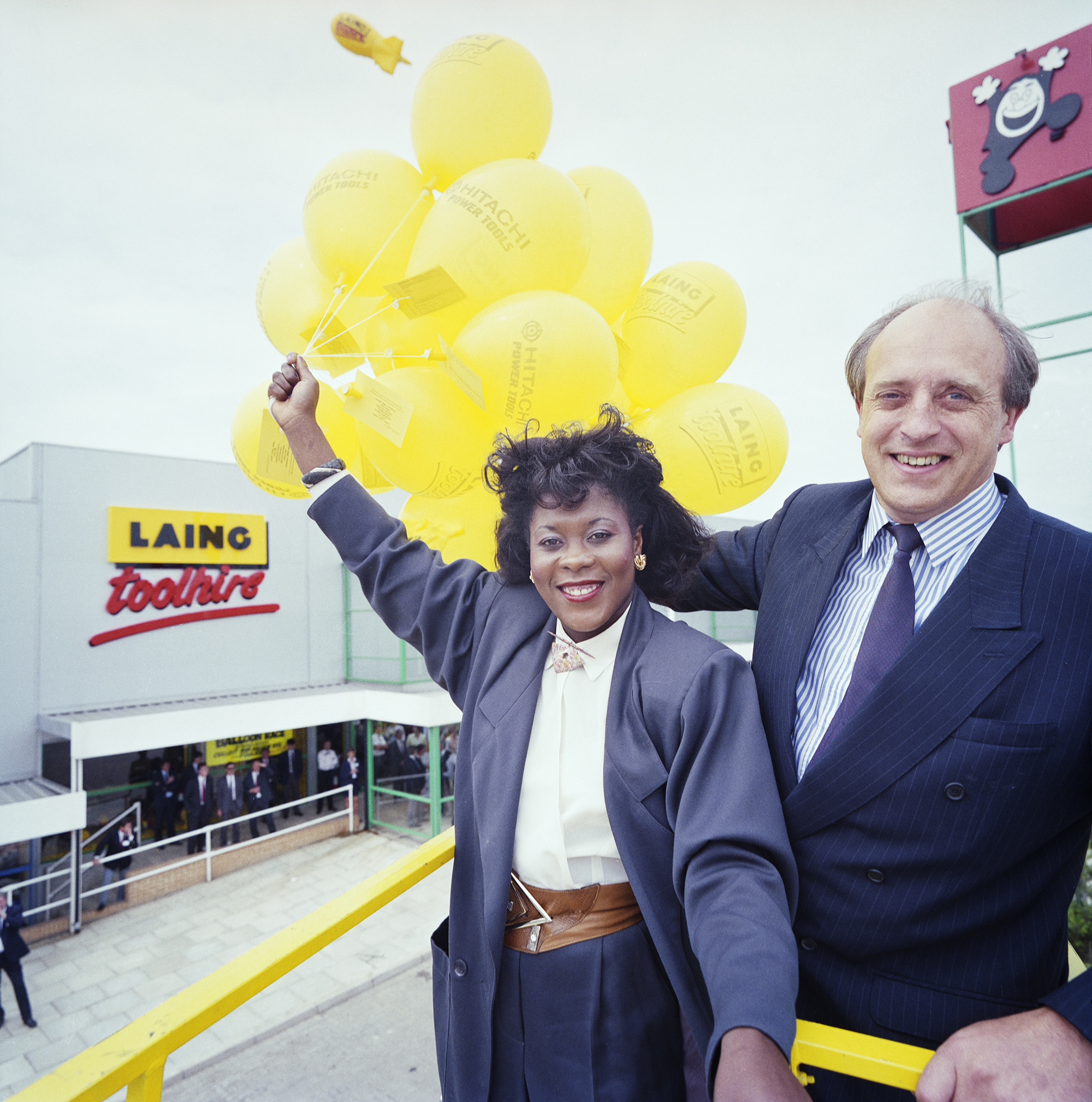 A man and woman pose for a picture. The man is on the right and wears a blue business suit. The woman on the left wears a grey suit and holds yellow balloons. In the background, there is a grey building with the title ‘Laing Toolhire’ on it.