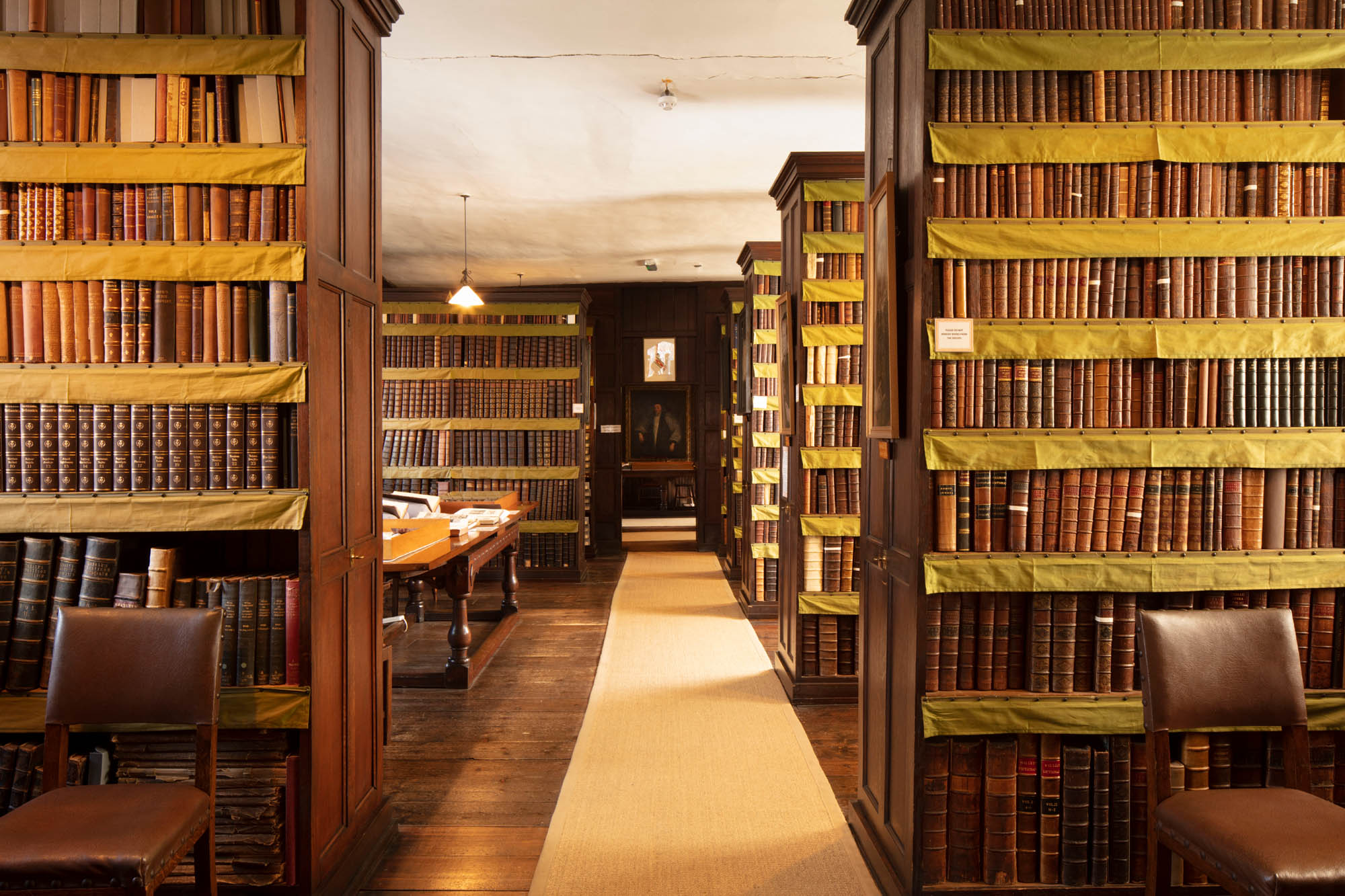 rows of dark wood historic bookcases line the Plume Library, filled with classic leatherbound books against a wooden timbered flooring lightened by a pale cream carpet runner.