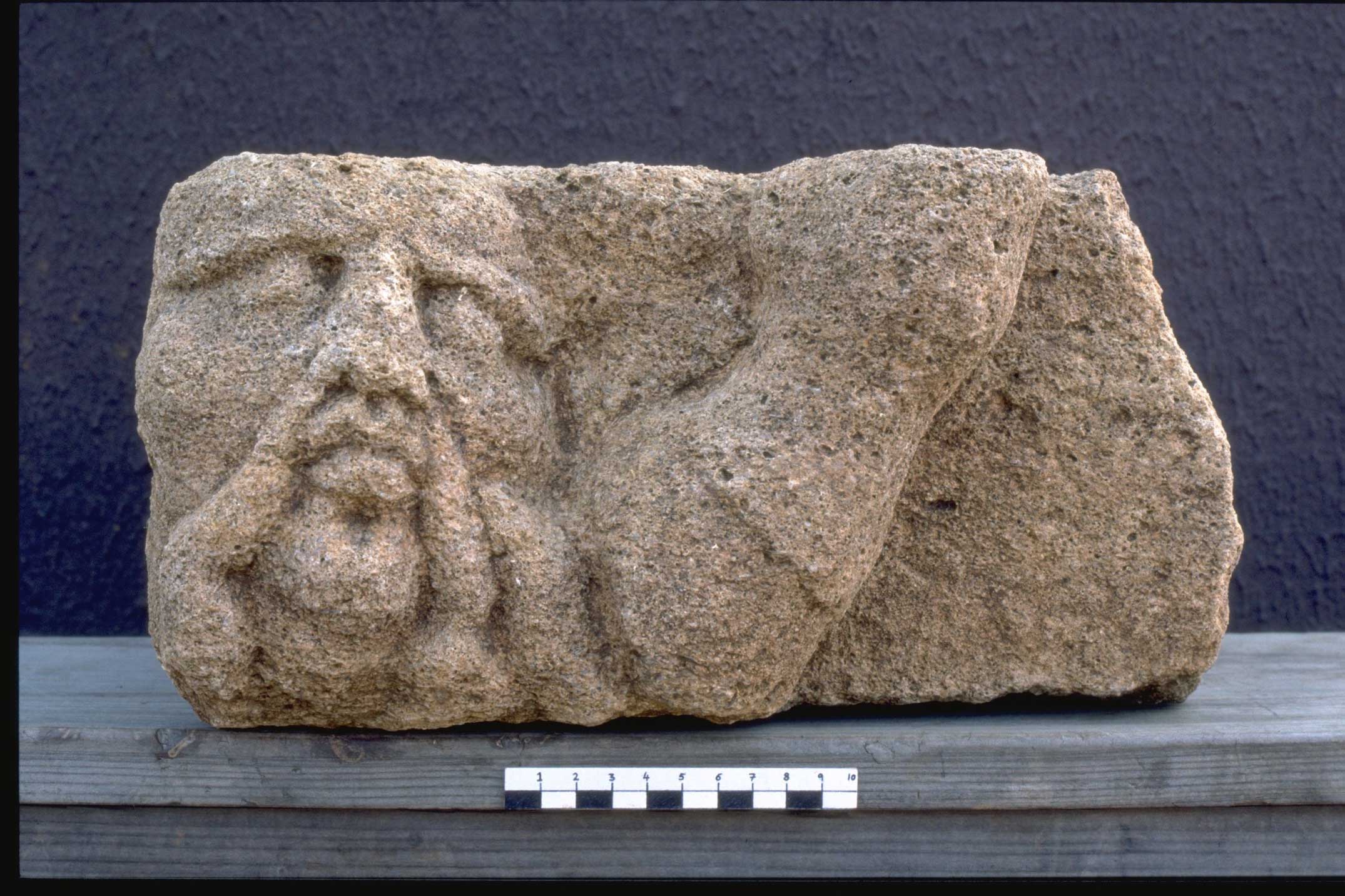 A fragment of stone sculpture depicting the head of a man with a long moustache being trampled by a horses hoof.