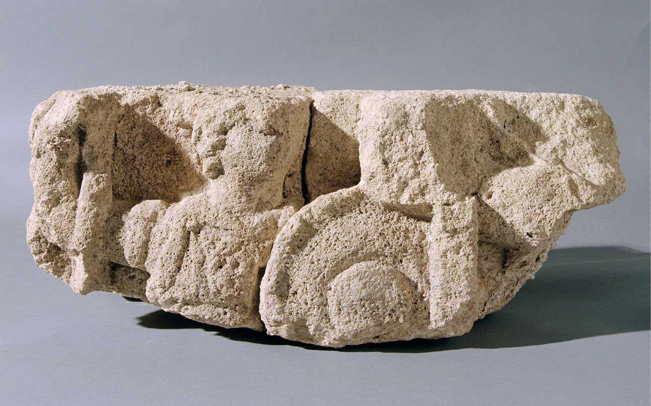 A fragment of stone sculpture depicting the upper part of a female figure carrying a spear and shield, with an arm of another  figure adjacent.