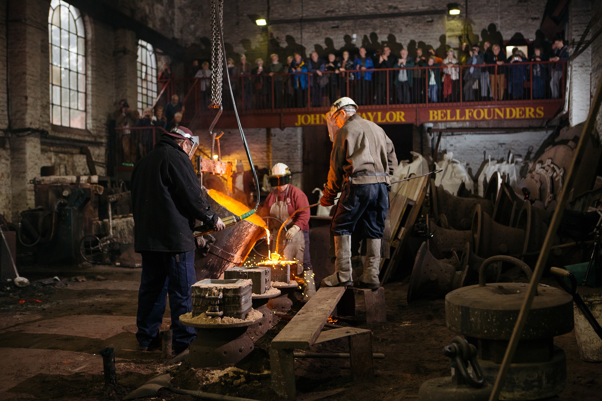 Three men pouring molten metal, demonstrating the bell casting process to a group of onlookers behind.