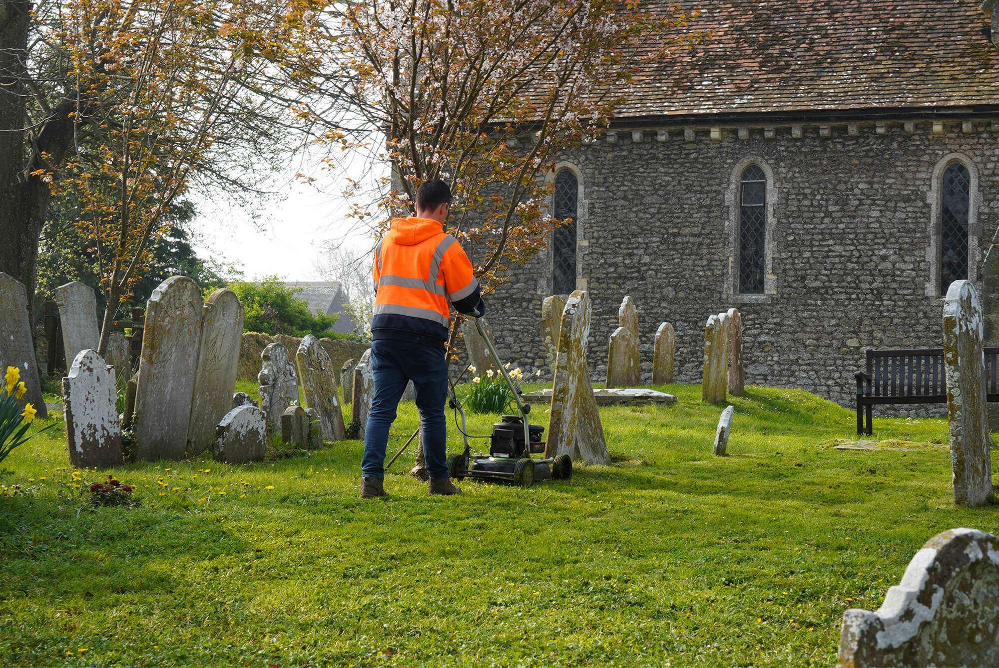Person wearing high-visibility jacket mowing grass in a grave yard, with grave stones around and part of the church in the background