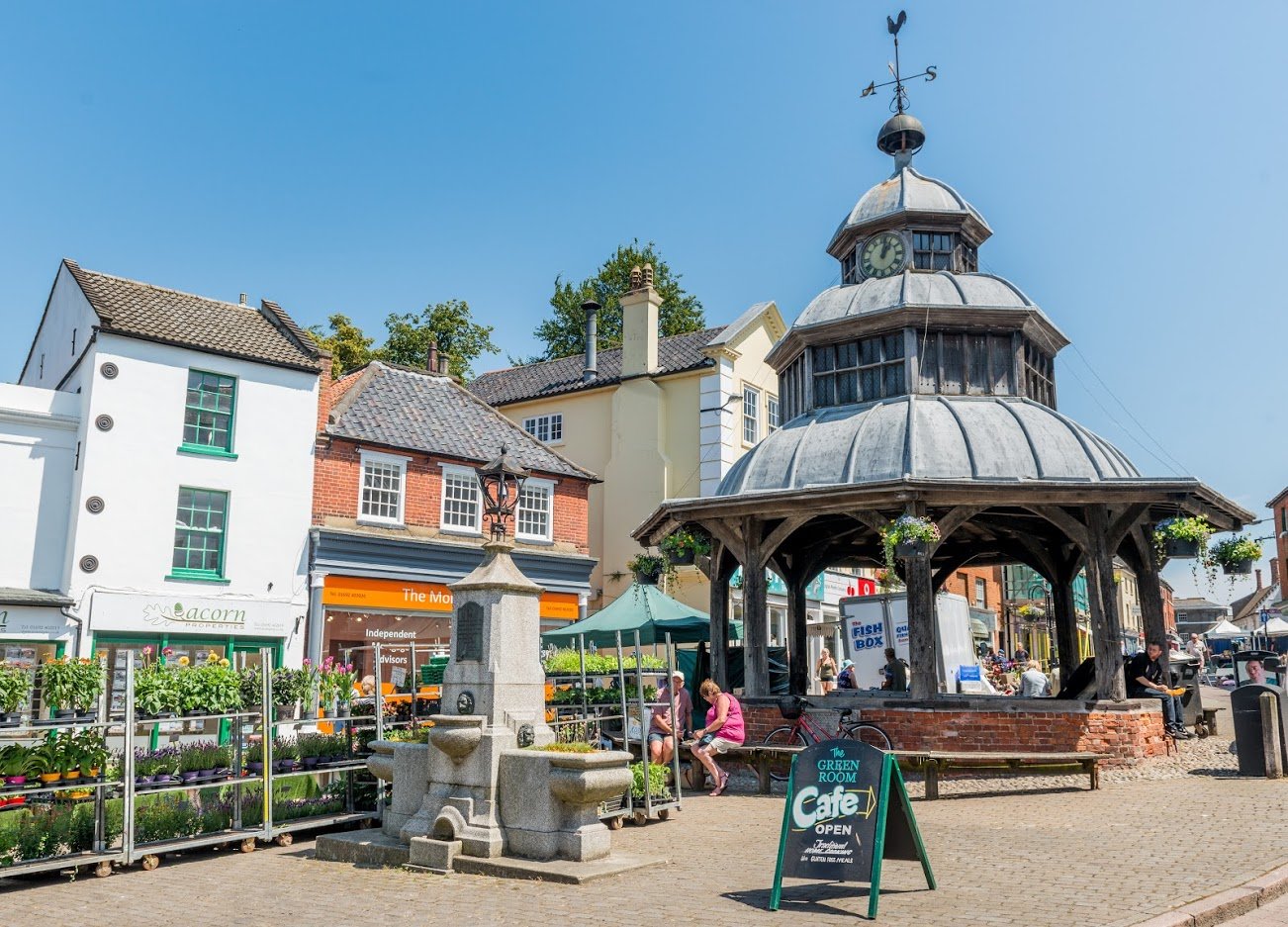 A market place with a market cross covered with a cupola, with historic buildings in use as shops in the background.
