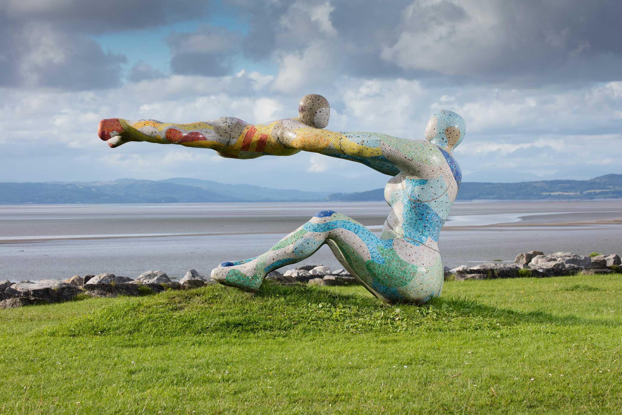 A sculptural installation in a coastal bay setting, depicting a seated female figure swinging a child in mid-air.