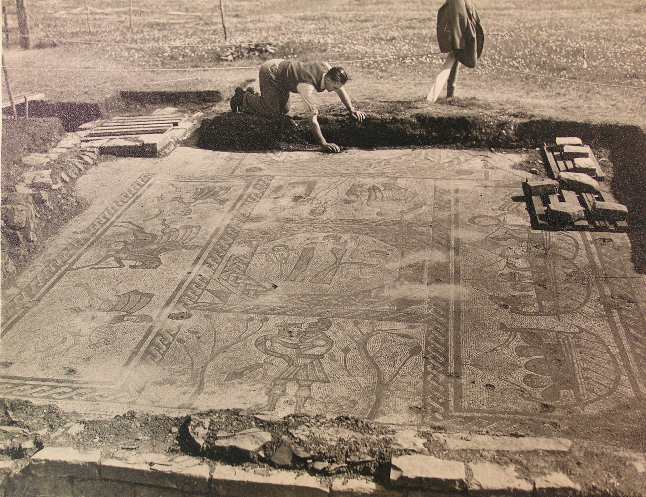 Black and white archive photograph of an archaeologist uncovering a buried mosaic floor depicting a classical mythological scene.
