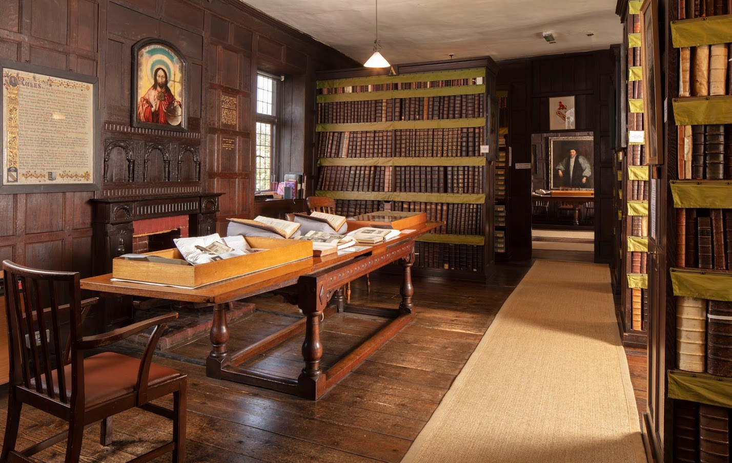 The library room shows dark wood panelling, wooden floorboard and dark wood bookcases with a long table to the left, covered in historic books. Historic portraits line the walls.