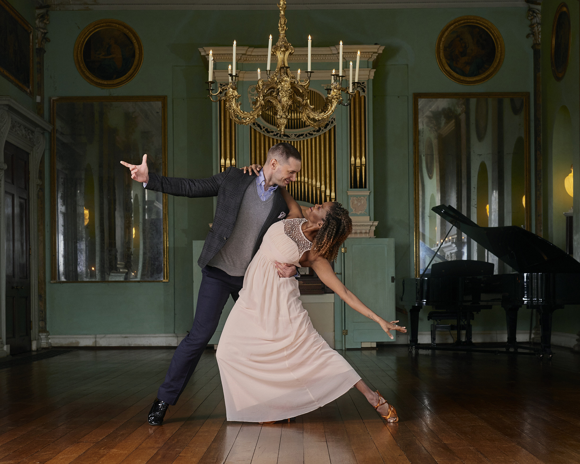 A couple dancing under a chandelier and a piano and organ in the background