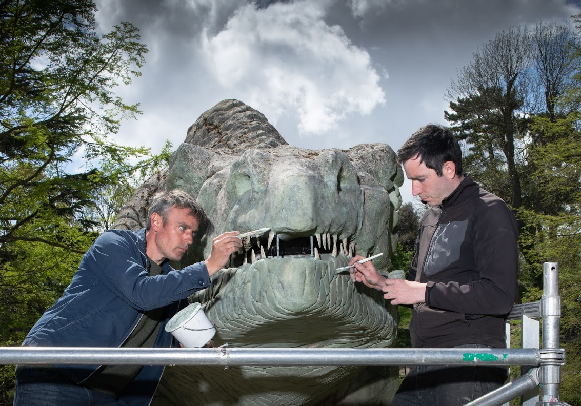 Photograph of the dinosaur sculpture's face as two skilled craftsmen intricately paint it's replacement jaw green.