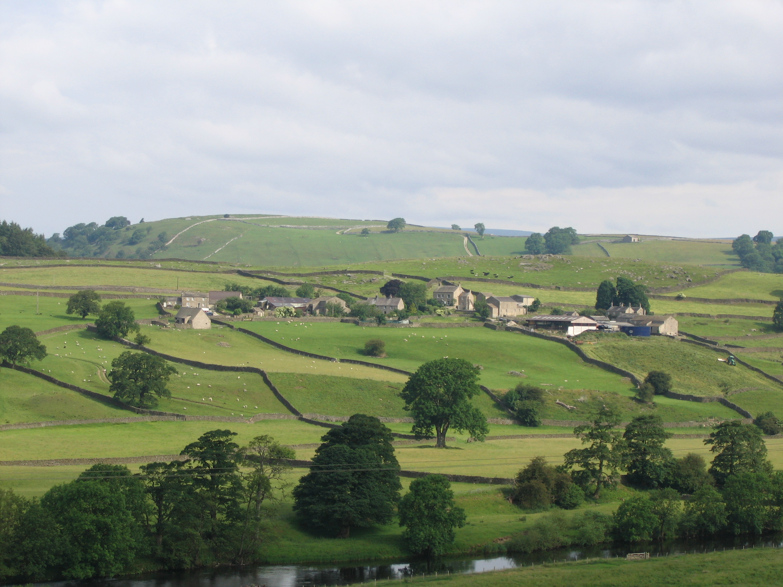 Village of Drebley in Yorkshire Dales National Park viewed from the SE across a river in the foreground.